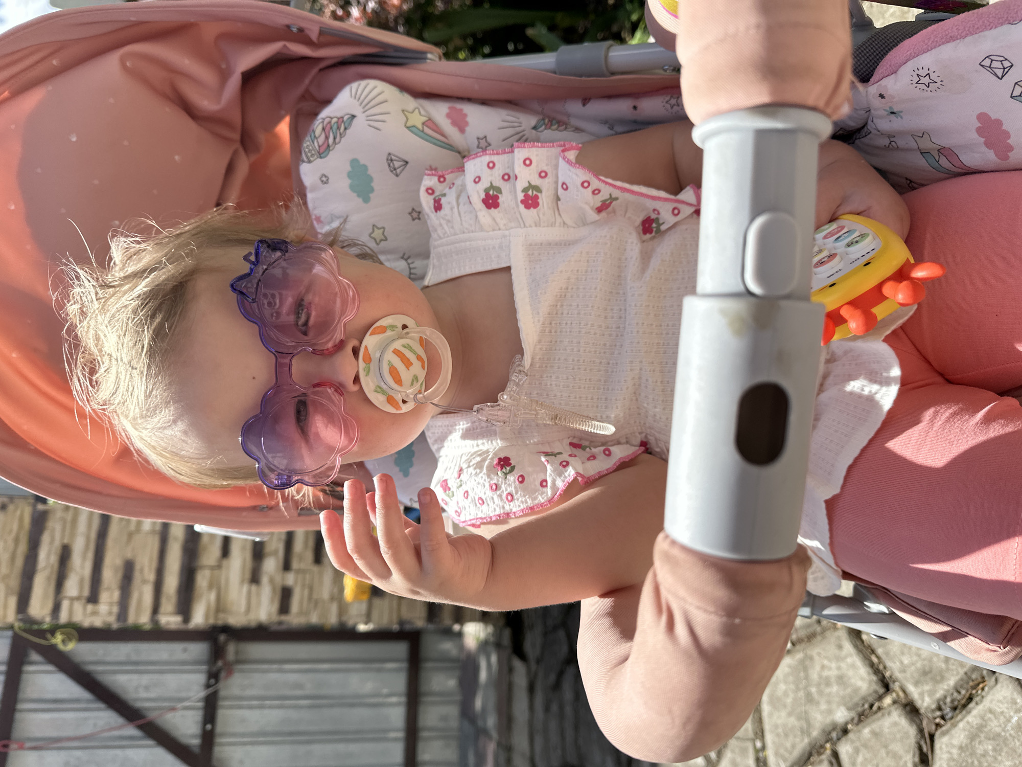 An adorable baby or toddler is captured in a close-up, angled shot, resting in a pink stroller under bright sunlight. The child has fair, wispy blonde hair and is wearing whimsical purple, flower-shaped sunglasses. A pacifier, adorned with small carrot illustrations, is in the child's mouth, attached by a clear clip to their clothing.
The baby is dressed in a white top with a textured pattern, featuring ruffled short sleeves detailed with pink stitching and embroidered red floral motifs. Pink trousers complete the outfit. The child's right hand is gently curled, resting near their body. In the other hand, the baby holds a vibrant yellow and orange toy, possibly a musical device or rattle, with four colorful circular buttons visible on its surface. The number "12.00" is faintly visible on the toy.
The interior of the stroller is padded with a white pillow decorated with playful patterns including unicorns, stars, clouds, and diamonds. The stroller itself is a soft pink color, and a gray handle or frame component is visible in the foreground. The setting appears to be outdoors on a sunny day, with strong shadows suggesting ample daylight. In the background, hints of a wooden fence or structure and paving stones can be seen on the ground, suggesting a garden or outdoor area in Nizhny Novgorod, Russia. The overall impression is one of a relaxed, sunny moment for the child.