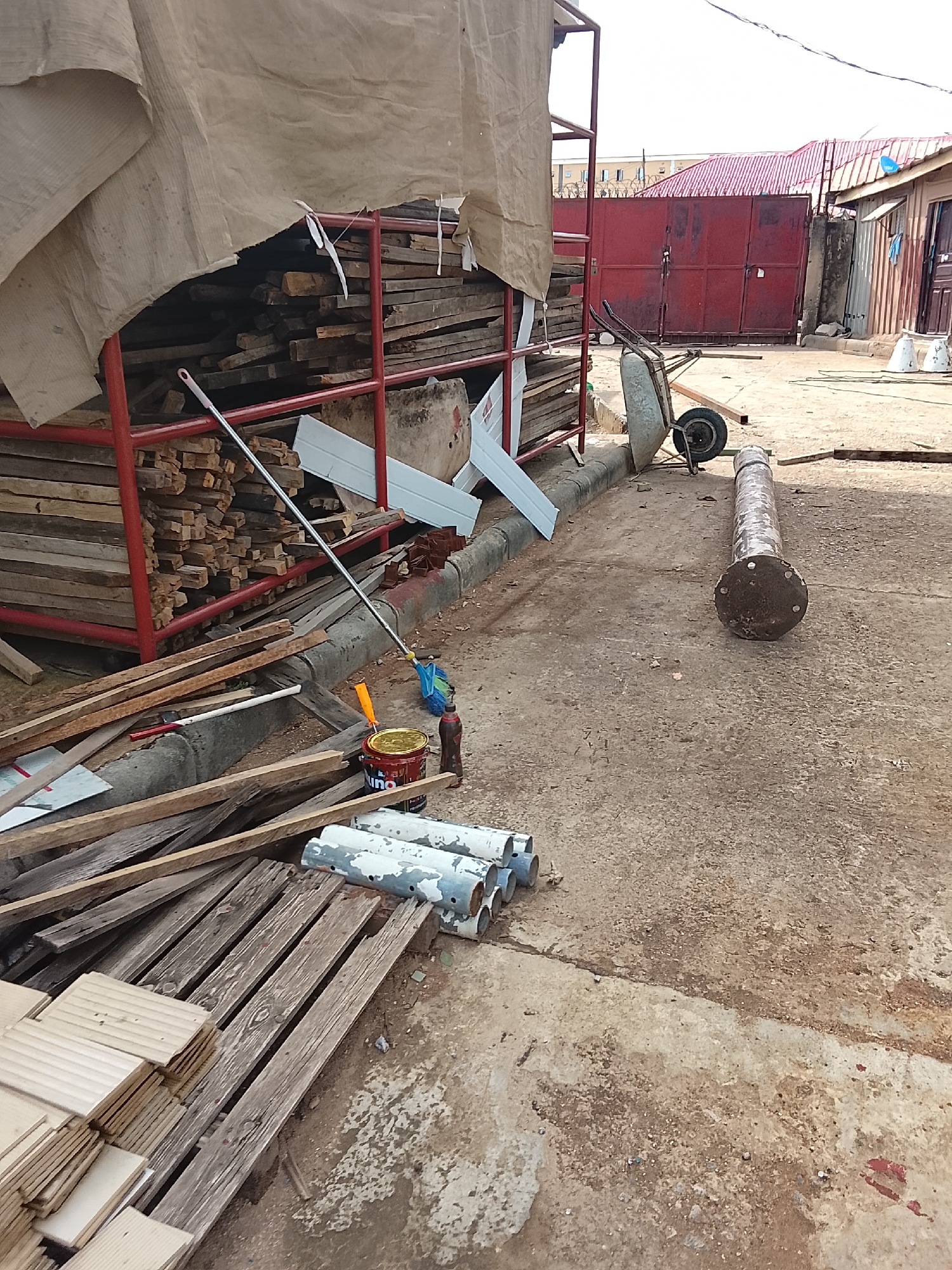 The image depicts a construction or renovation site in Abuja, Nigeria. The foreground is cluttered with building materials including stacks of lumber, wooden planks, white painted pipes, and metal sheeting. A red metal rack filled with lumber is covered with a tan tarp. A painting setup with a can of paint, brushes, and thinners sits amidst the materials. A large, heavy, cylindrical metal pipe with a flanged end lies on the concrete pavement. In the background, a red metal gate and a building with a red roof are visible. A wheelbarrow is nearby, suggesting ongoing work. The ground is concrete, stained and weathered. The scene is sunlit. There are no people visible.