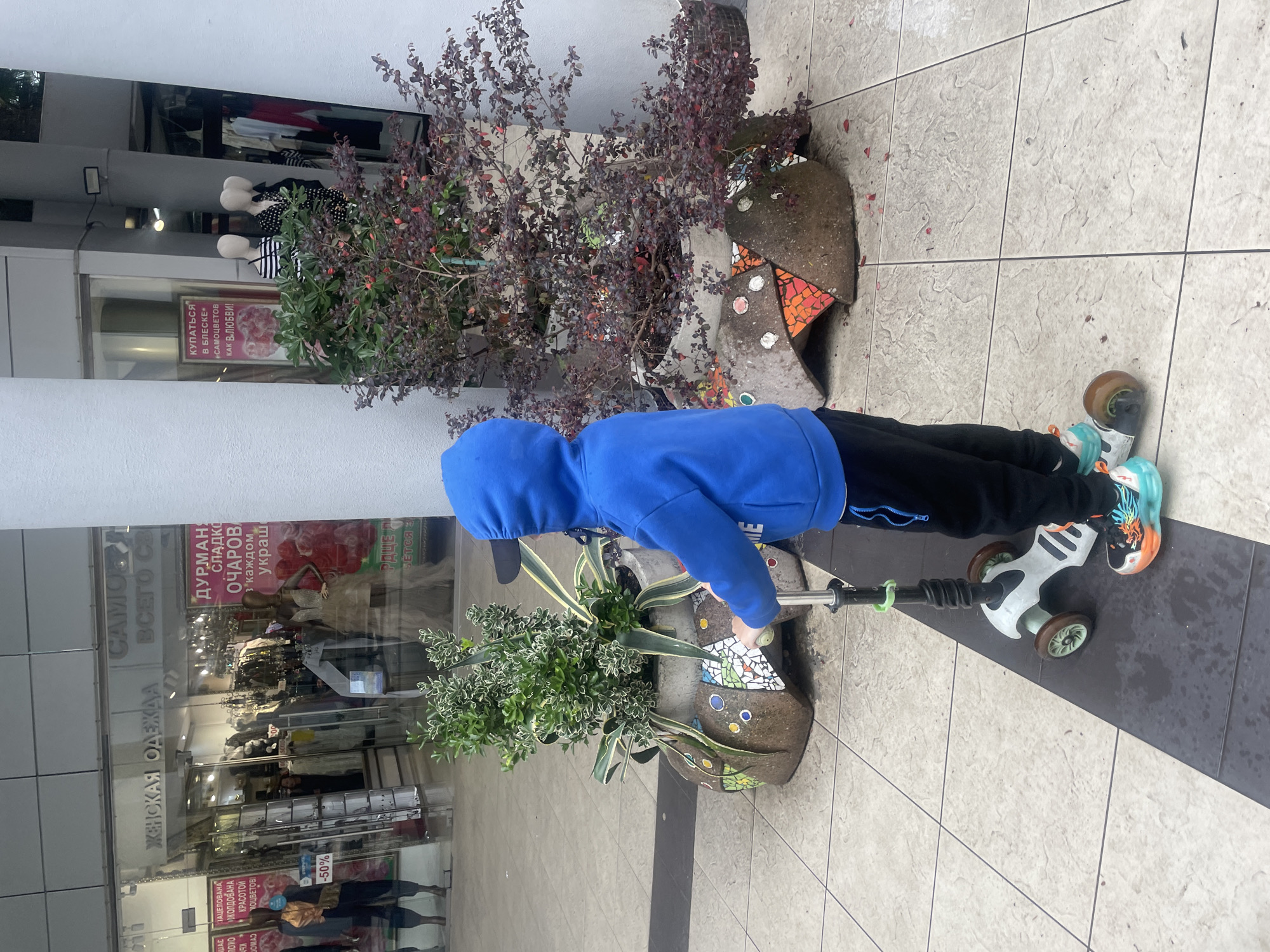 A child in a blue hooded sweatshirt and black pants is standing on a white scooter with three wheels. The child is on a tiled surface in front of a shop. There are two large potted plants on either side of the child. The shop behind the child is a women's clothing store. The sign on the shop is written in Russian. The OCR text in Russian includes references to Sochi ("SAMO").