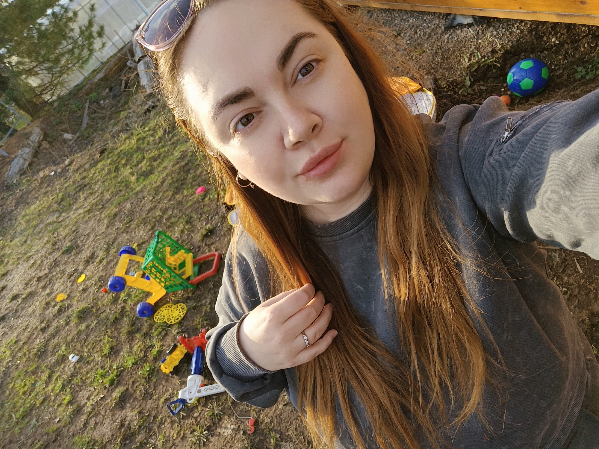 A medium shot captures a woman with light skin, auburn hair, and sunglasses perched atop her head. She's wearing a gray sweatshirt and a ring on her left hand. The background consists of a patch of earth covered with sparse grass and scattered children's toys, including a shopping cart, building blocks, and a soccer ball. A greenhouse structure is visible in the upper left corner of the image. The location is outdoors, potentially a backyard or play area. There is no clear evidence of a specific location within the city of Chaikovsky, Russia.