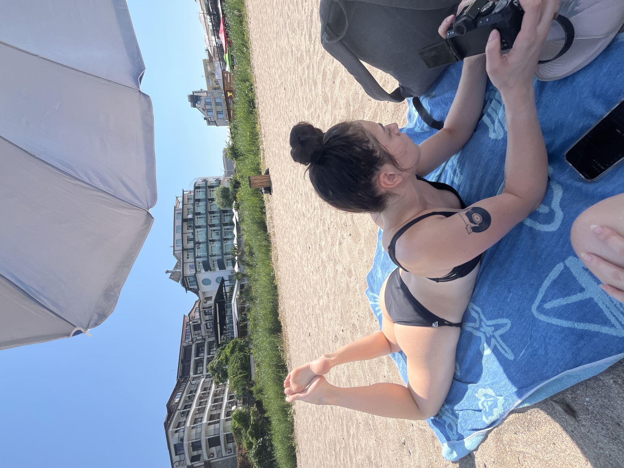 A young woman is lying on her stomach on a blue towel on a sandy beach in Lozenets, Bulgaria. She is wearing a black bikini. Her hair is pulled up in a bun. She is looking at a camera that she is holding in her hands. To her right is a grey backpack. In the background are several multi-story buildings and some green vegetation. The sky is clear and blue. A large white umbrella is partially covering the top of the image.