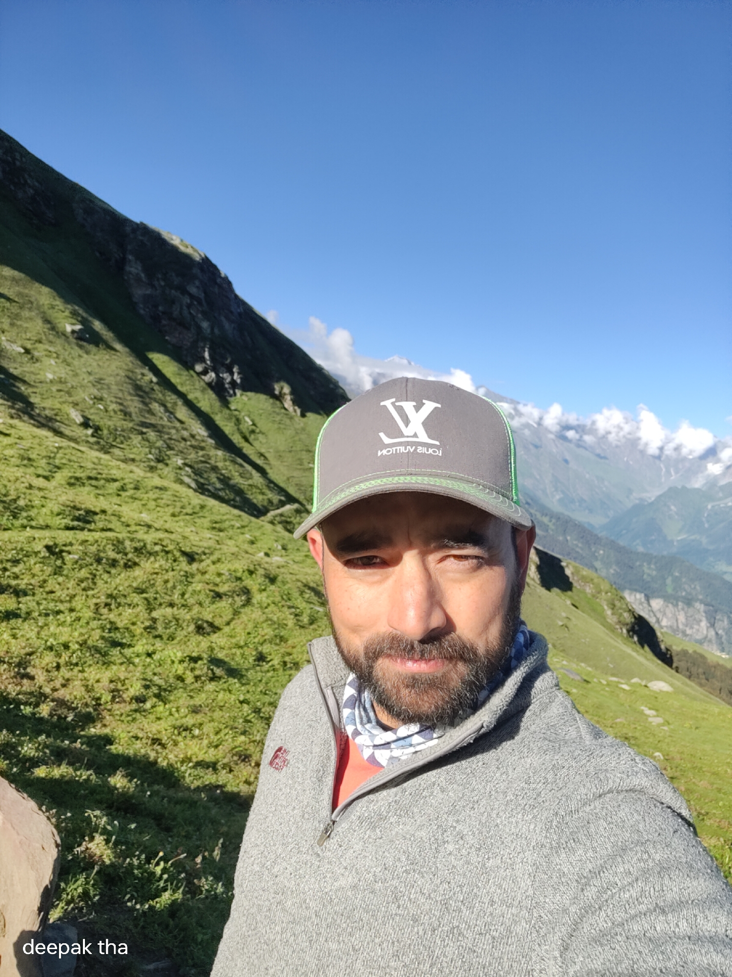 The image is a selfie taken by a man in the mountains, likely in or near the city of Bashisht, India, given the presence of the text "deepak tha" in the lower left corner, which could be a watermark or caption. The man is wearing a grey sweater with a red t-shirt underneath, a patterned neck gaiter, and a grey baseball cap with a neon green brim and a logo that appears to be "LV". He has a beard and is looking at the camera. The background is a steep, green, grassy mountainside under a clear blue sky. Distant mountains, some with snow-capped peaks and clouds, are visible in the background, suggesting a high-altitude location. The sun is casting shadows on the grassy slopes, indicating it is daytime.