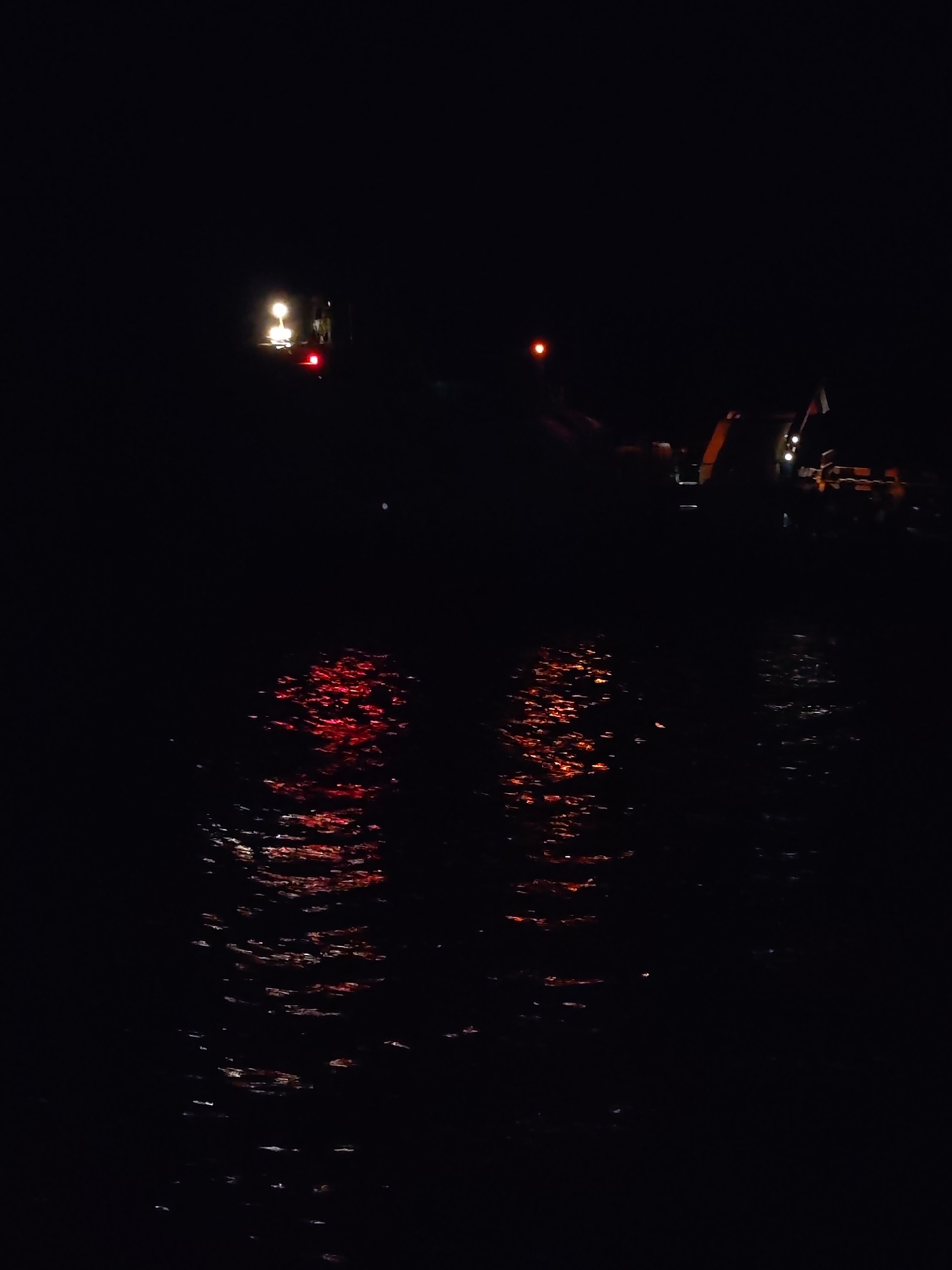 The image shows a very dark scene, likely at night, with reflective water in the foreground. The reflections are primarily red. In the upper part of the image, there are faint lights suggesting the presence of machinery or vehicles. On the left, two lights are visible, the one at the top looks like a floodlight and has a red glow right below it. On the right, a taller structure can be seen with faint white lights, possibly an excavator or crane along with a collection of objects beside it. The environment is generally dark and indistinct. The location is Unknown, Unknown.