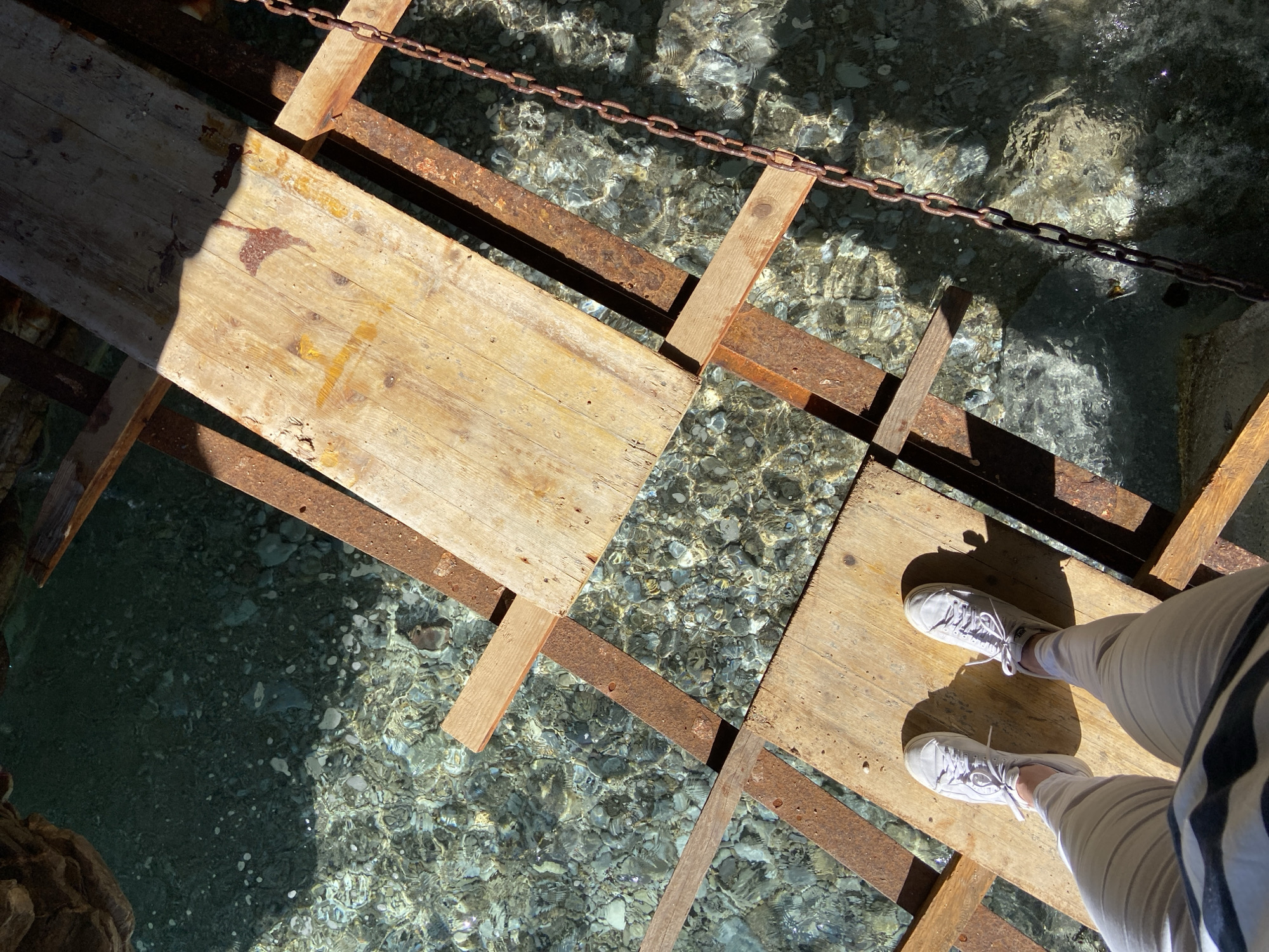 A top-down, angled shot showcases a rustic wooden bridge over a clear, rocky stream in Sveti Stefan, Montenegro. The bridge's construction features weathered wooden planks supported by rusty metal beams and wooden struts. A rusty chain runs parallel to the bridge's edge. Sunlight penetrates the water, creating shimmering reflections on the stream bed. A person stands on the bridge, their feet visible, clad in white sneakers and light-colored pants. The person is wearing a striped top. A shaded rocky bank is visible in the lower left corner.