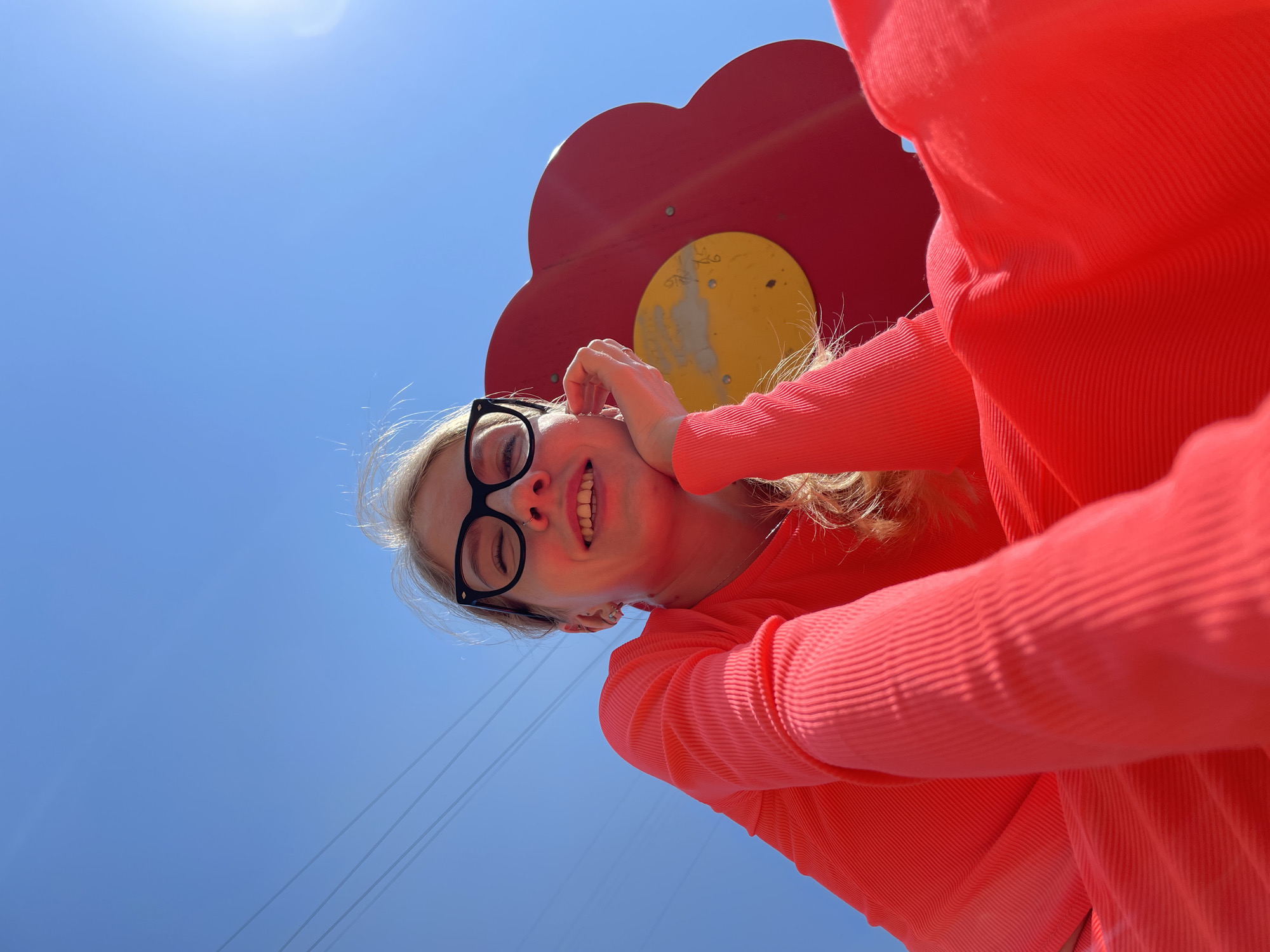 A low-angle shot captures a smiling woman with blonde hair and glasses, wearing a red shirt, seemingly positioned beneath a playground structure with a red flower-shaped element and a yellow center. The clear blue sky fills the background. Thin cables stretch from the upper left corner of the frame. It is not possible to determine the specific location in Izhevsk, Russia based on the information provided.