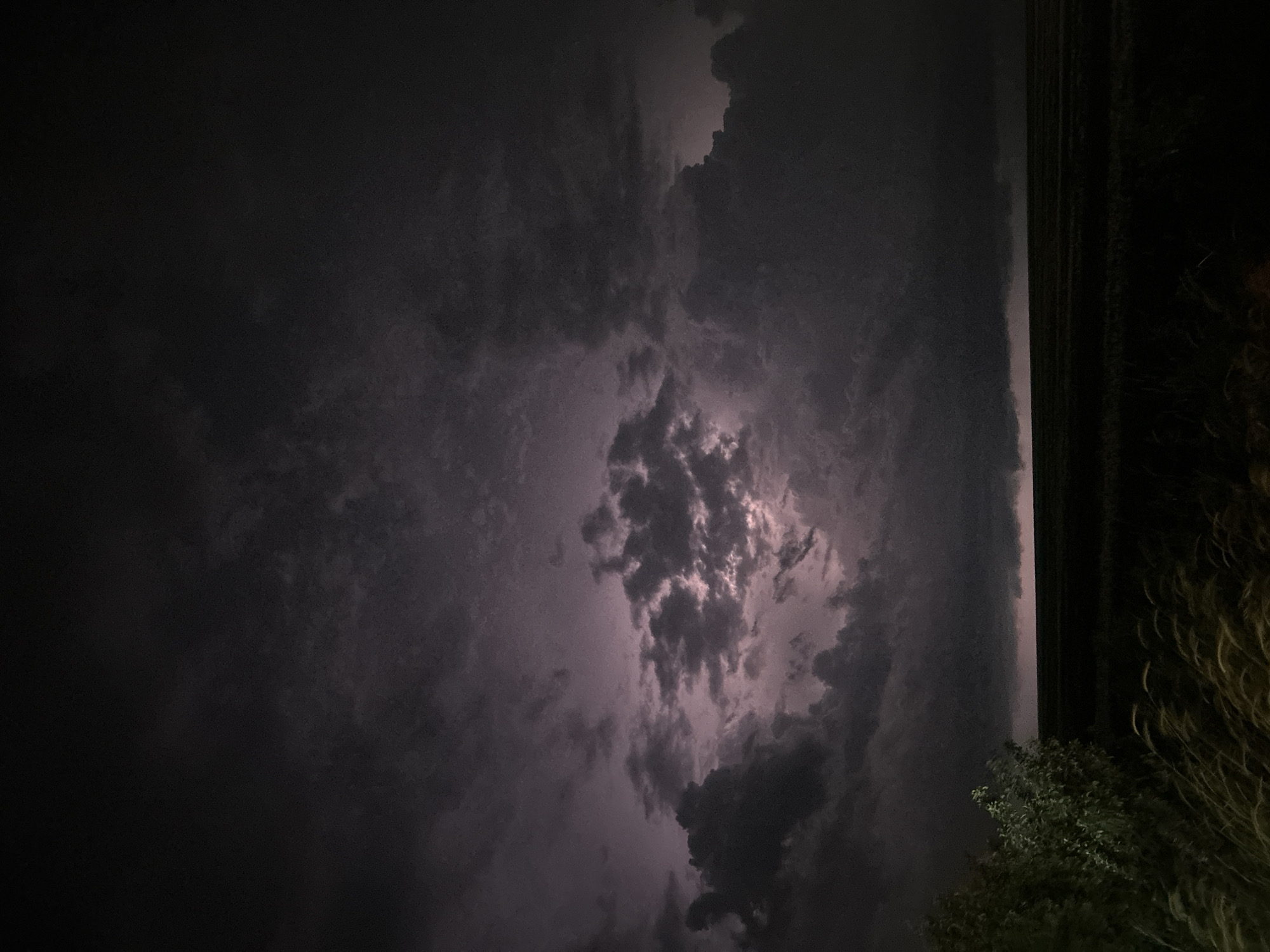 A nighttime, low-angle view shows a dark sky filled with dense, textured clouds. A bright area illuminates the center of the cloud mass, potentially caused by lightning. To the right, a dark, vertical structure, possibly a building edge, is visible, accompanied by foliage. The location and the actions and interactions are not able to be determined.