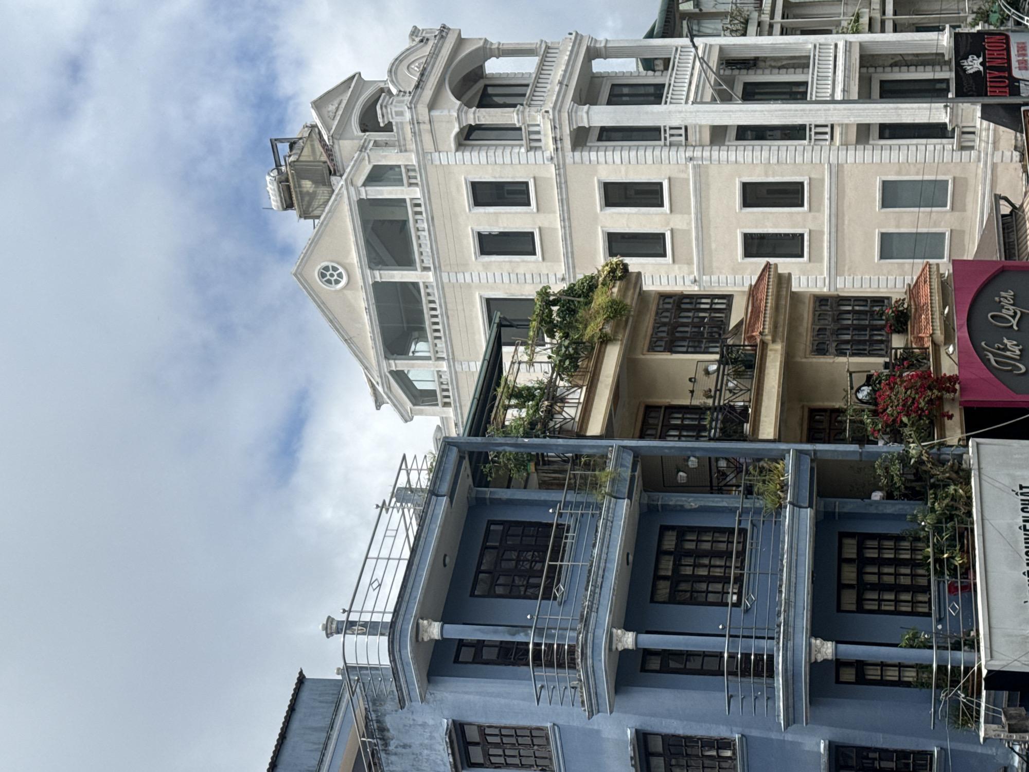 The image depicts a street scene in Da Lat, Vietnam, showcasing a row of multi-story buildings. The primary focus is on a prominent light blue building on the left, featuring several balconies with metal railings and dark-framed windows. Adjacent to it is a cream-colored building with more elaborate architectural details, including columns, arches, and balconies. Further to the right, another building with a lighter facade and similar balcony structures is visible. Red flowers cascade down from a balcony on a building between the blue and cream structures. The sky above is overcast with some visible clouds. Signs with Vietnamese text are present on the lower levels of the buildings. No people or specific actions are discernible in the image. The environment appears to be an urban setting.