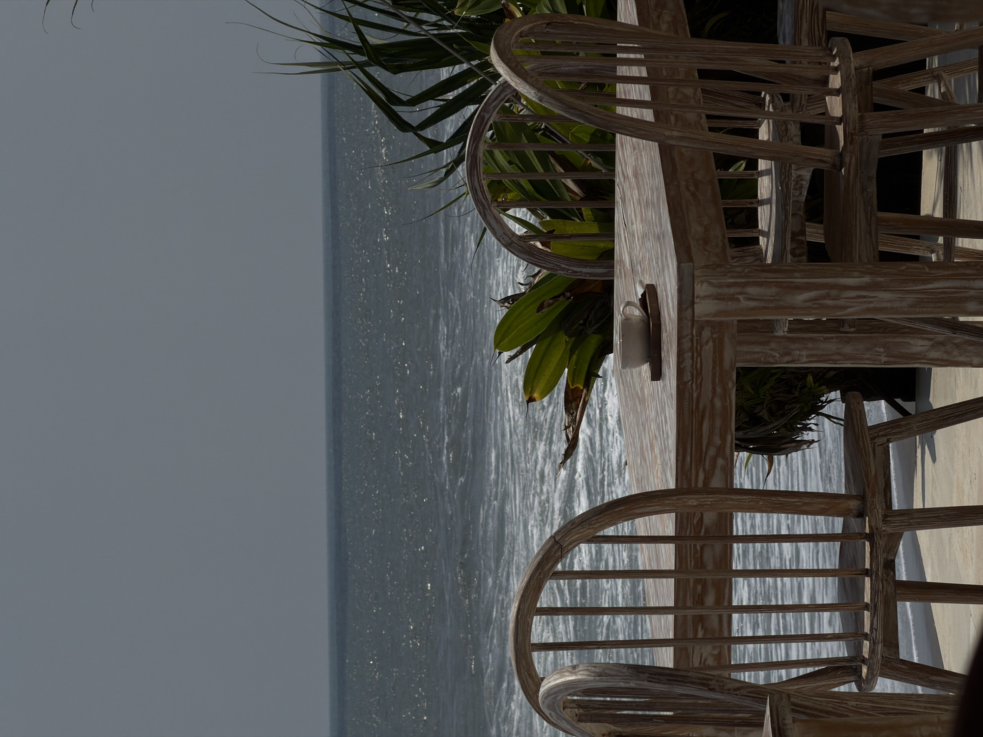 An outdoor seating area in Belalang, Indonesia, features a weathered, light-colored wooden rectangular table with four matching spindle-back chairs, two prominently facing the ocean. A white ceramic cup rests on a dark brown coaster on the table surface. Beyond the seating, a dark blue-gray ocean with white-capped waves occupies the mid-ground, extending to a hazy gray horizon beneath an overcast sky. Lush, spiky green tropical foliage partially frames the ocean view from the right and center-right. The ground beneath the table and chairs is a light-colored, flat surface. No individuals are present in the scene.