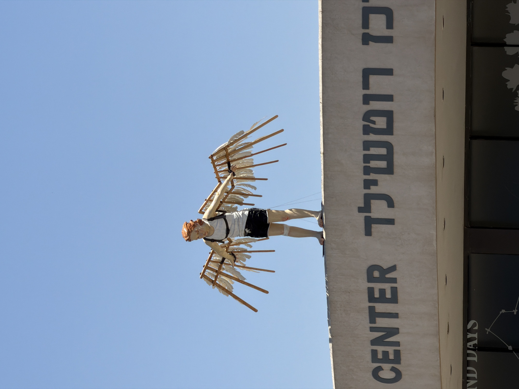 A sculpture depicting a human-like figure stands atop a light-colored, flat-roofed building against a clear blue sky. The figure has reddish-orange hair, pale skin, and is dressed in a white sleeveless tank top and black athletic shorts with white stripes. It wears a harness on its back, to which two large, rudimentary wings are attached. The wings are constructed from wooden frames with white feather-like elements. The figure is barefoot and appears to be in a stance suggestive of preparing for flight or having just landed, with its arms slightly outstretched and gaze directed downwards.
The building's facade below the sculpture features large, dark, sans-serif letters. On the left, the English word "CENTER" is visible. To its right, Hebrew characters read "רוטשילד", which transliterates to "Rothschild". This identifies the location as the Rothschild Center, situated on Rothschild Boulevard in Tel-Aviv, Israel. Below the main facade, a darker section with windows reveals partial text "ND DAYS" on the far left. The scene is brightly lit by natural sunlight, indicating daytime.