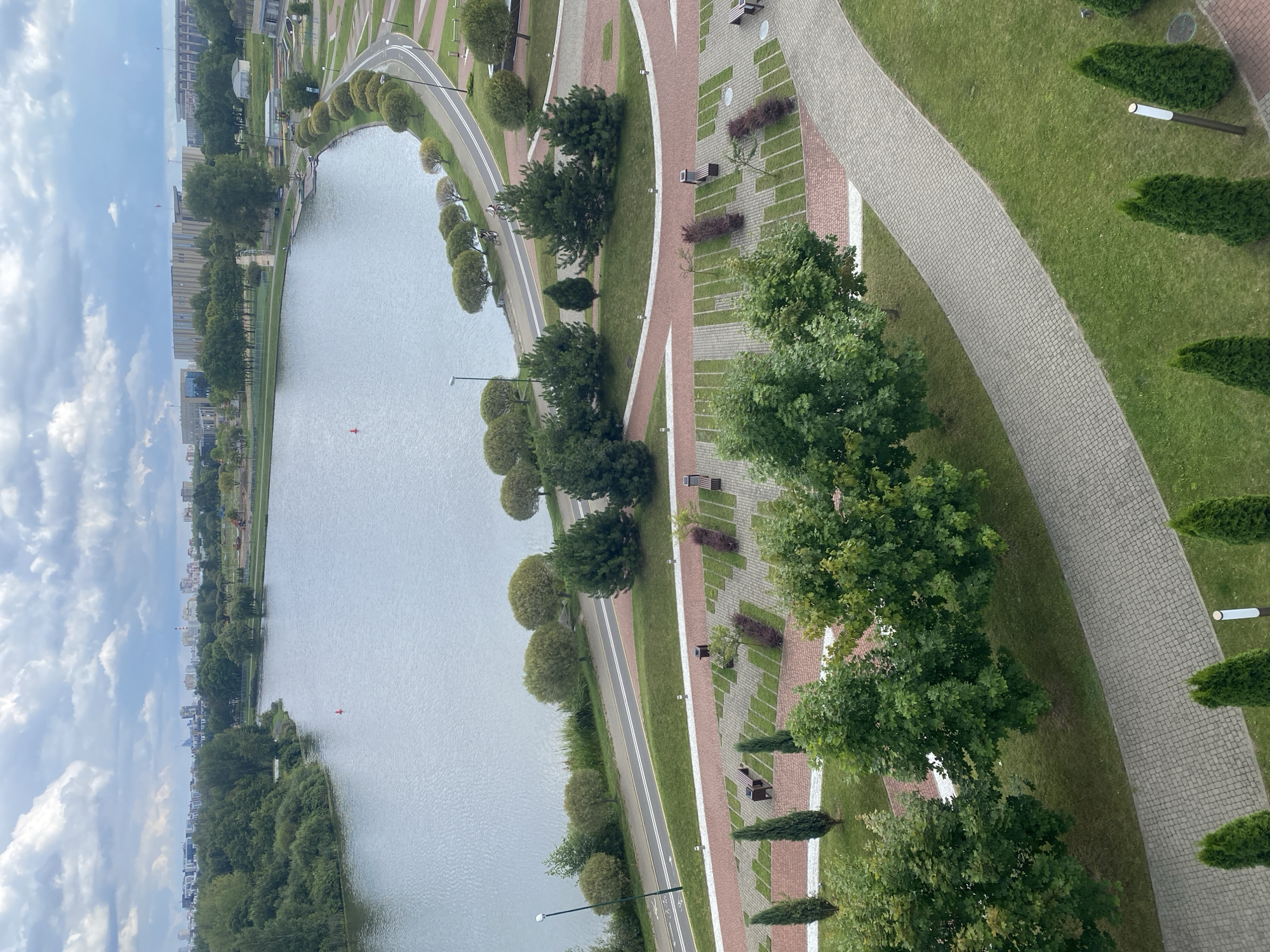 This is an aerial photograph of a park area with a lake in Minsk, Belarus. The scene captures a large, calm body of water on the left side of the image, bordered by lush green trees and vegetation. A paved pathway with white lane markings for bicycles and pedestrians curves along the edge of the lake. In the foreground, there is a paved parking area with designated spots and several benches. Green grass and a variety of trees, including evergreen and deciduous types, are scattered throughout the park. In the distance, urban buildings and more trees are visible under a cloudy sky. There are no people clearly identifiable in the image, though two cyclists can be seen on the pathway near the lake.