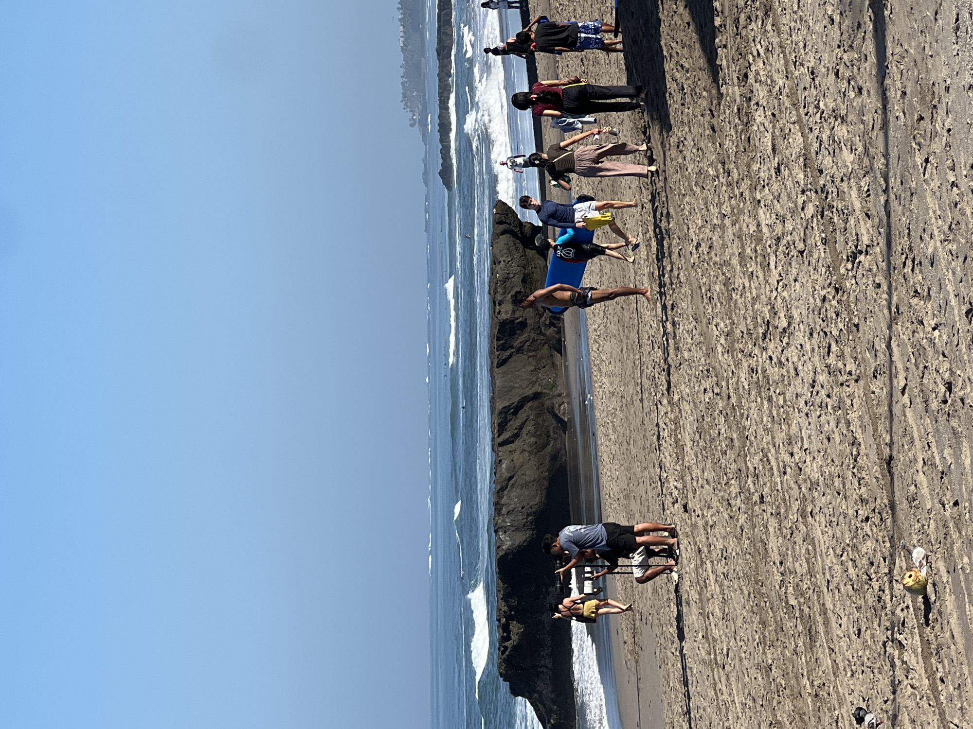 The image depicts a sunny day at a beach in North Kuta, Indonesia. In the foreground, a sandy beach is textured with tire tracks. A small, round object with a brown and yellow pattern rests on the sand. A group of people are scattered across the beach.
In the mid-ground, to the left, two individuals are operating a video camera on a tripod, focusing on a woman walking away from them towards the ocean. She is wearing a yellow swimsuit. Further to the right, a group of people are gathered, some carrying a blue surfboard. Another person is standing with a surfboard.
The background features the ocean with moderate waves and white surf. A large, dark rock formation juts out of the water. In the distance, the coastline is visible with scattered buildings and palm trees. The sky is clear and blue.