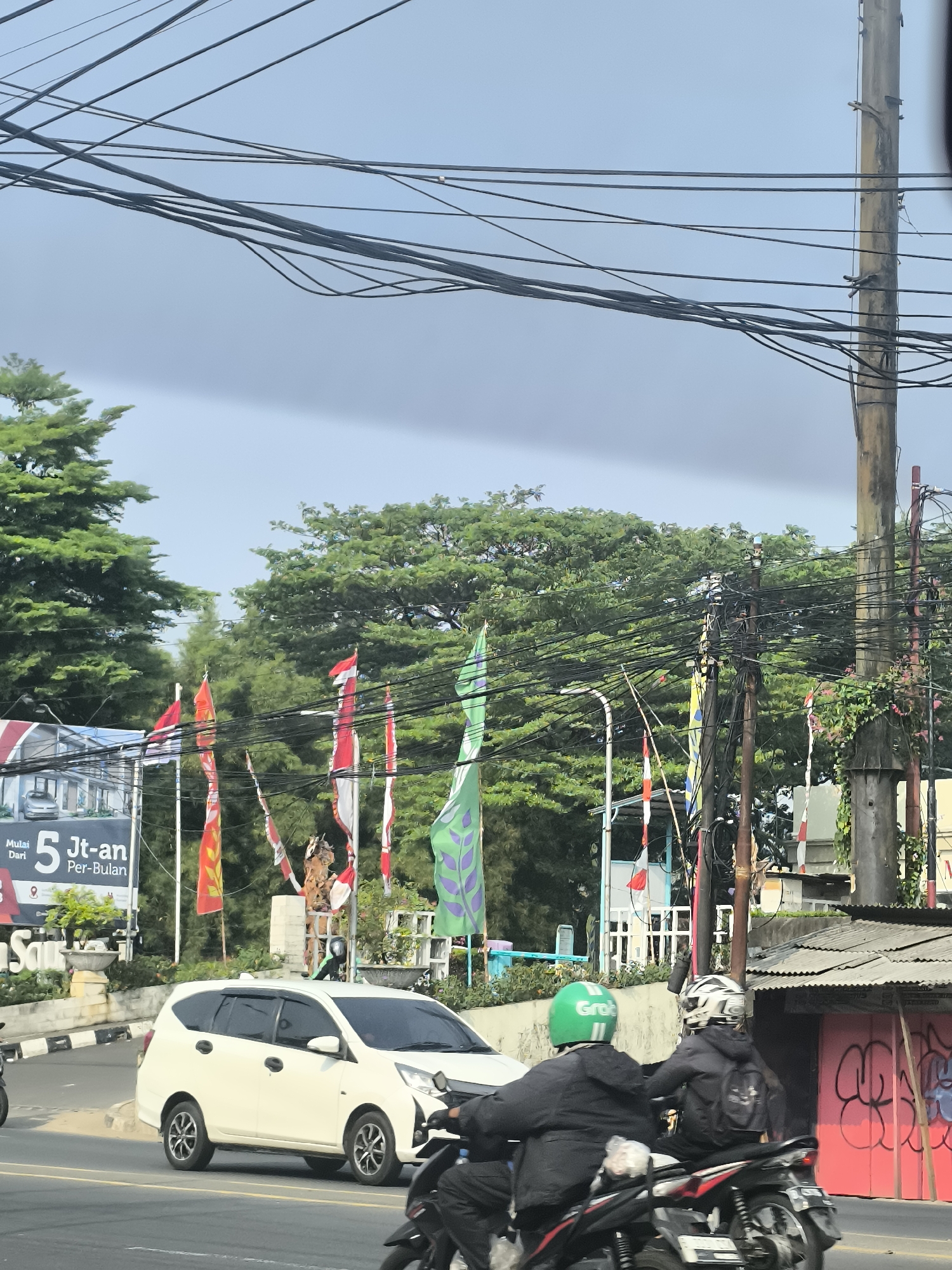 The image depicts a street scene in Tangerang, Indonesia, on an overcast day. The foreground is dominated by a white minivan, likely a Toyota Avanza, driving on a paved road. Behind the minivan, two individuals on a motorcycle are visible, with the rider wearing a green helmet and the passenger a white and black helmet.
A variety of flags are displayed along the roadside, including Indonesian flags (red and white) and green flags with a leaf-like design. These flags are attached to poles and appear to be part of a street decoration or event.
In the background, lush green trees form a dense canopy. Several utility poles with a complex network of wires are a prominent feature of the urban environment. A small, weathered building with a corrugated metal roof and red painted doors covered in graffiti is also visible on the right side of the frame. To the left, a billboard with text indicating a price and "Per-Bulan" (per month) is partially visible. The overall impression is a typical street view in a Southeast Asian city, characterized by its traffic, vibrant street decorations, and abundant vegetation.