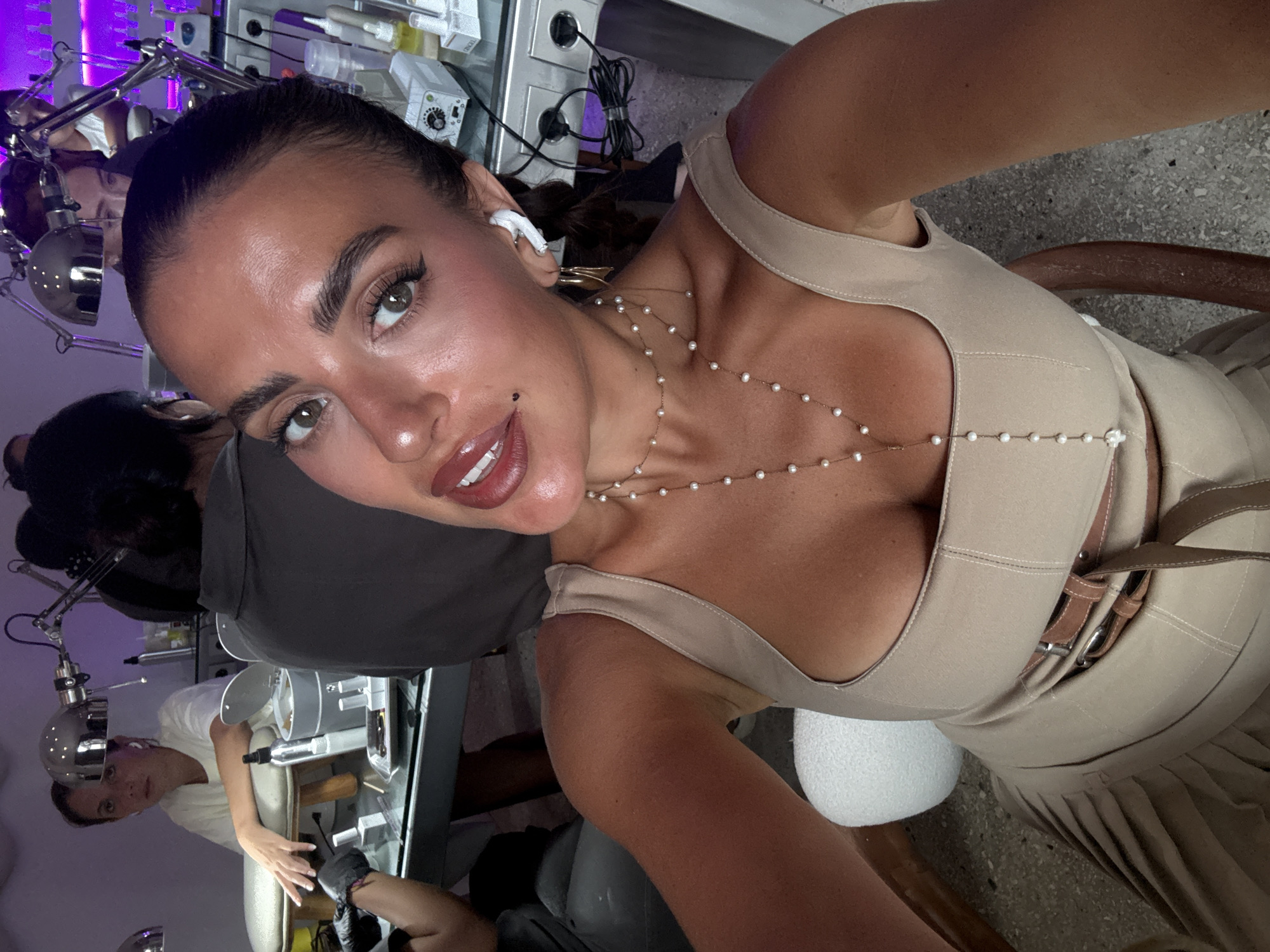 A tan-skinned woman with dark hair pulled back is taking a selfie in what appears to be a nail salon. She's wearing a light tan dress with a plunging neckline and a double-buckled belt. A delicate pearl necklace adorns her neck. She has a mole on her face, is wearing eyeliner, and her lips are glossy. In the background, other individuals are present, one wearing a grey t-shirt and another wearing a white t-shirt. There are several pieces of equipment consistent with nail care stations, including adjustable lamps, power outlets, and tools. Based on the lighting and equipment present, the location is likely in a beauty or personal care setting. The location in the city of North Kuta, Indonesia, is assumed from context clues of popular destinations for tourism.