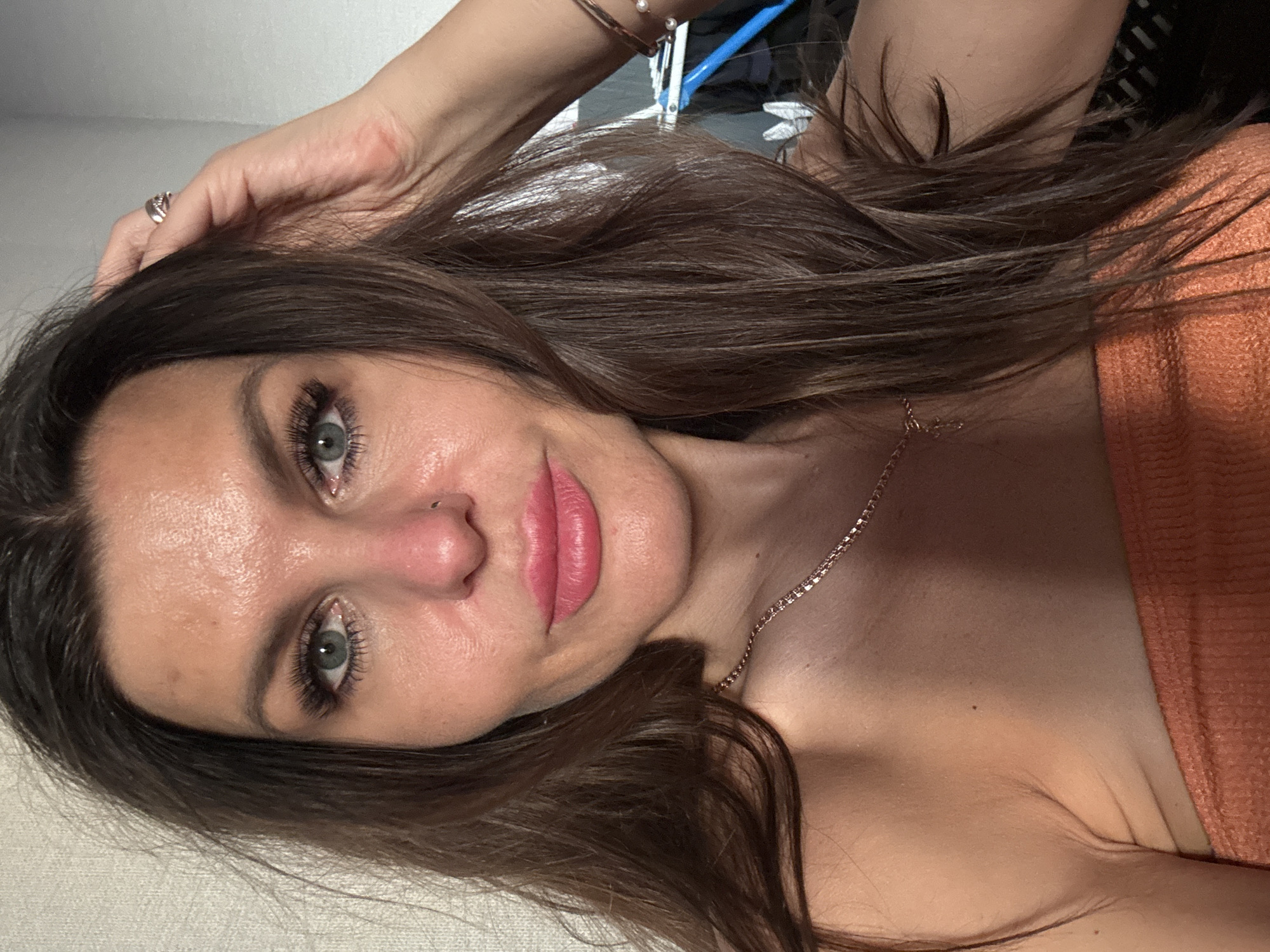 A close-up, vertically oriented portrait of a woman with long, dark brown hair. She has fair skin, green eyes, and is wearing pink lipstick. A gold chain necklace is visible on her neck. Her right hand is raised towards her hair. She is wearing an orange garment on her right shoulder. The background appears to be a white or light gray wall. The location of the city of Chaikovsky, Russia is not discernible from the visual information provided.