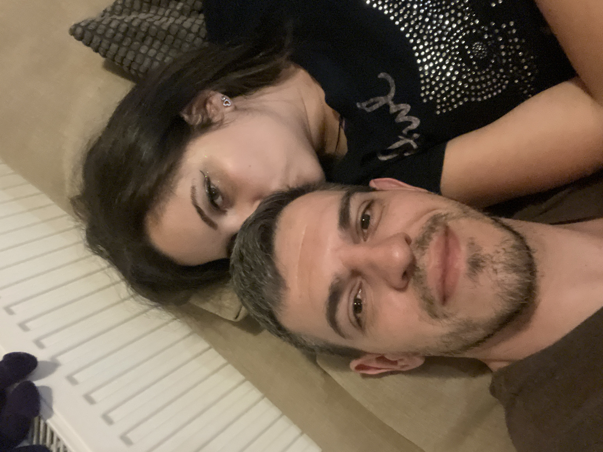The image captures a close-up, high-angle selfie of a couple. The man, in the foreground on the left, has short, greying hair and a beard, and is wearing a brown t-shirt. He is looking directly at the camera. The woman is leaning in from the right, kissing the man on the forehead. She has long, dark hair and is wearing a black t-shirt adorned with rhinestones. Her left ear is visible, featuring a small heart-shaped earring. They are positioned on what appears to be a couch, with a beige sofa and a patterned cushion visible. In the background, a white radiator with vertical bars is present, and what looks like a dark blue plush toy is resting on top of it. The setting suggests an indoor, domestic environment. There are no discernible elements that indicate the specific location within the city of Sofia, Bulgaria.