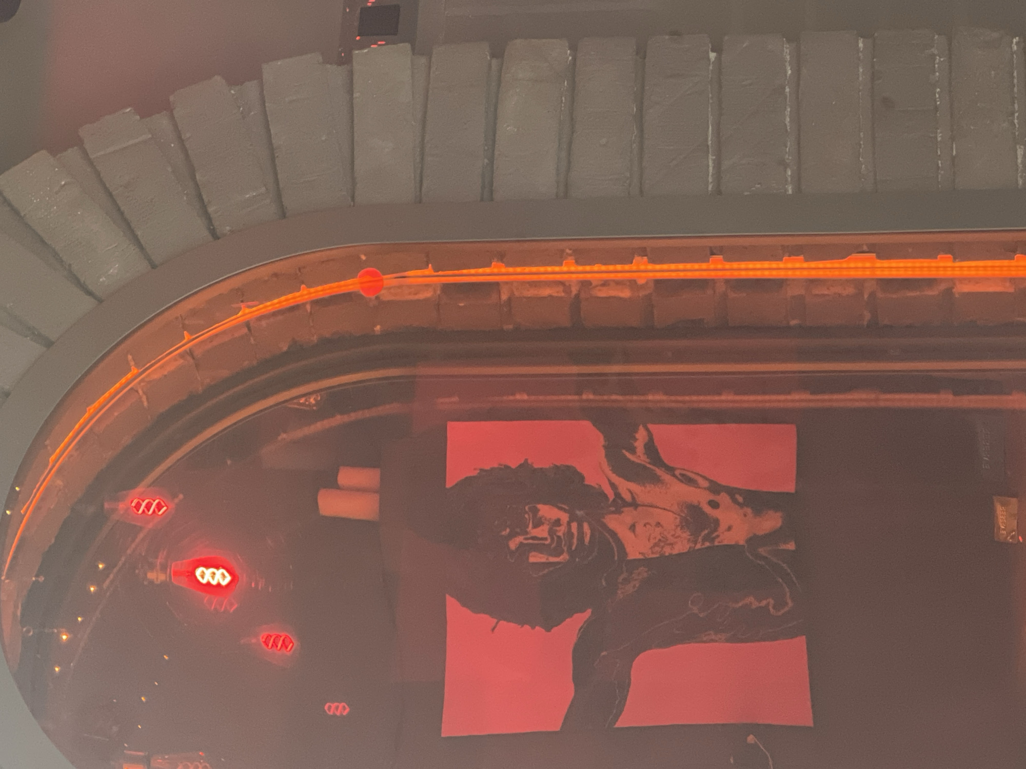 The image depicts an interior scene, possibly a bar or restaurant, with a prominent arched doorway. To the left, within the archway, a red poster of a man with dark, wild hair and an open chest is visible. He appears to be spread-eagled, with his arms outstretched. Above the poster, several red Edison-style light bulbs are hanging. To the right of the poster, two unlit white candles stand on a dark surface.
The archway itself is outlined with a bright orange light strip. The exterior of the archway, to the right of the image, is constructed of grey, stacked rectangular blocks, giving it a textured, somewhat rough appearance. The overall lighting is dim and dominated by the red and orange hues from the bulbs and light strip. The exact location in Sofia, Bulgaria, is not discernible from the visual information alone.
