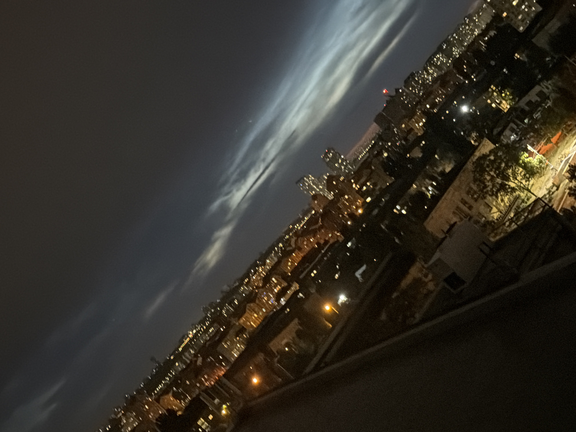 The image is a nighttime, high-angle shot of a city skyline, angled downwards from left to right. The sky is dark with streaks of light-colored clouds, possibly noctilucent clouds, indicating a clear night with high-altitude ice crystals. Below the sky, a dense urban landscape is visible, illuminated by numerous artificial lights from buildings and streetlights. The city appears to be Kyiv, Ukraine, based on the characteristic architecture and density of buildings. The perspective suggests the photo was taken from a high vantage point, such as a rooftop or a tall building.
In the foreground, on the far left, a dark, out-of-focus structure, likely a balcony or railing, occupies a significant portion of the frame, suggesting the viewpoint of the photographer. The mid-ground reveals a mix of apartment buildings and other structures with illuminated windows, creating a warm glow against the night sky. Some taller buildings with more concentrated lights are visible in the distance. The overall impression is one of a sprawling city at night, with a sense of depth and expanse. There are no visible people or vehicles in the frame, and the scene is static. The quality of the image is somewhat grainy, consistent with low-light photography.