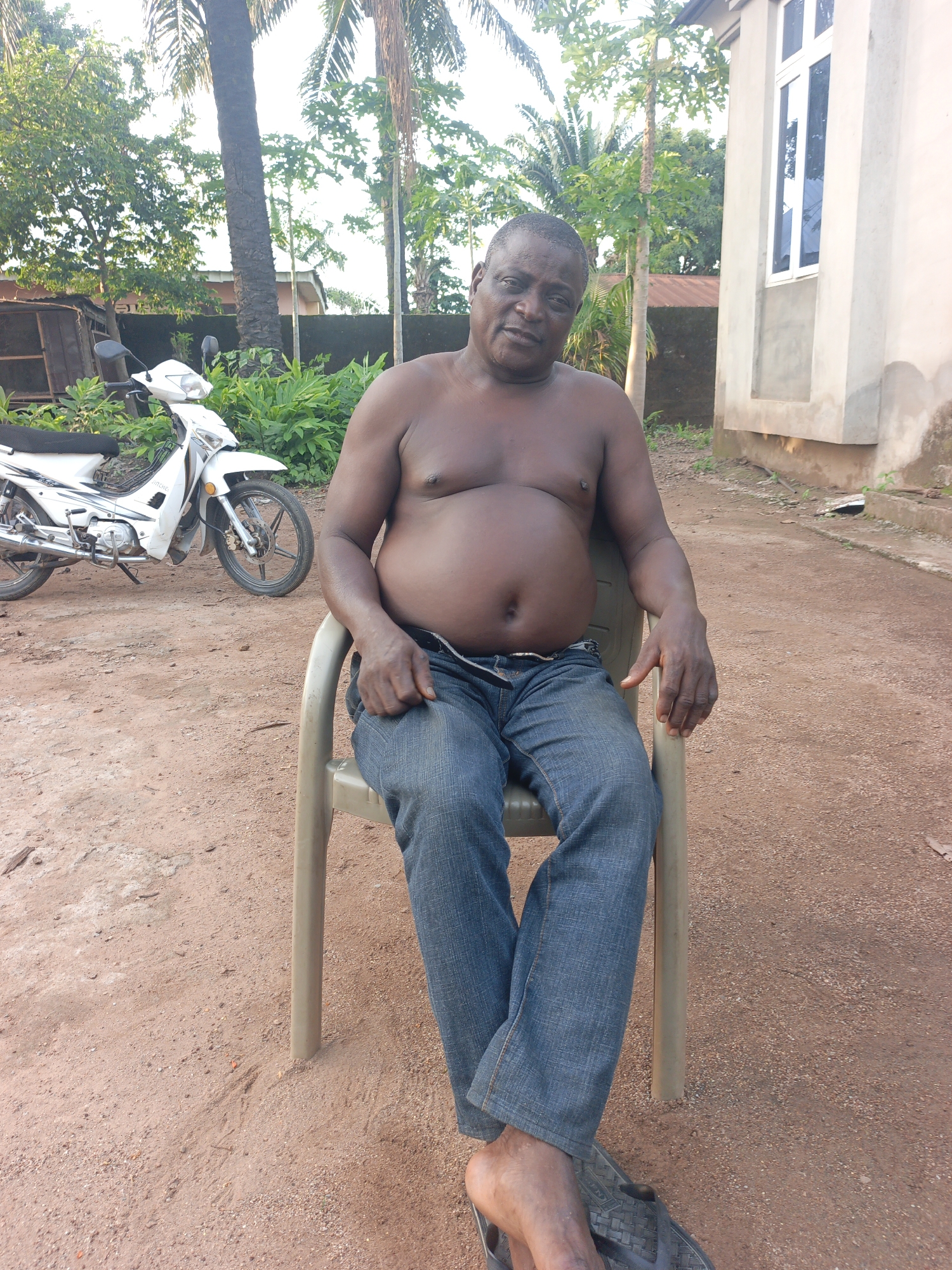 A medium, outdoor shot shows a shirtless man sitting on a plastic chair in Akwanga, Nigeria. He wears jeans and sandals. A white motorcycle is parked nearby. The ground is reddish-brown dirt. Trees and a building are in the background.