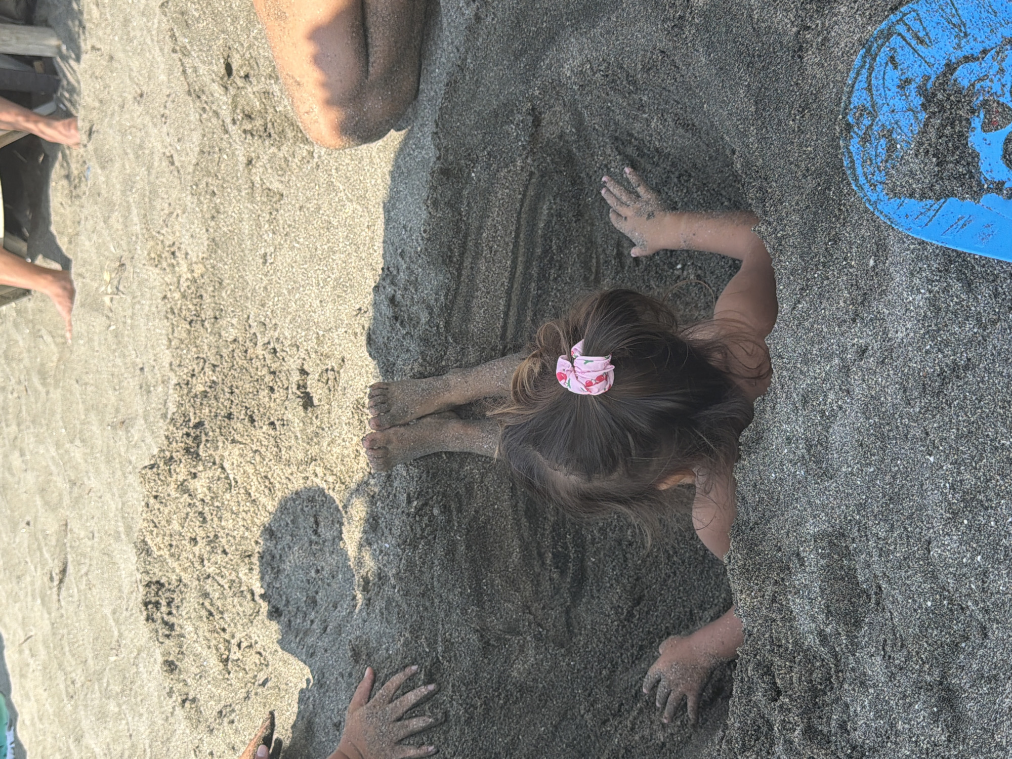 The image depicts a child buried in dark sand up to their shoulders on a beach. The child has brown hair styled with a pink scrunchie and is facing away from the camera. Their feet, covered in sand, are visible sticking out of the sand. To the left of the child, another set of sandy hands is visible. In the lower right corner of the image, a blue plastic object, possibly a toy or a beach accessory, is partially visible, also covered in sand. The background shows more sand and shadows, suggesting a sunny day. The scene is set on a beach, likely a public one, given the presence of other people's legs in the background. This beach is located in Burgas, Bulgaria.