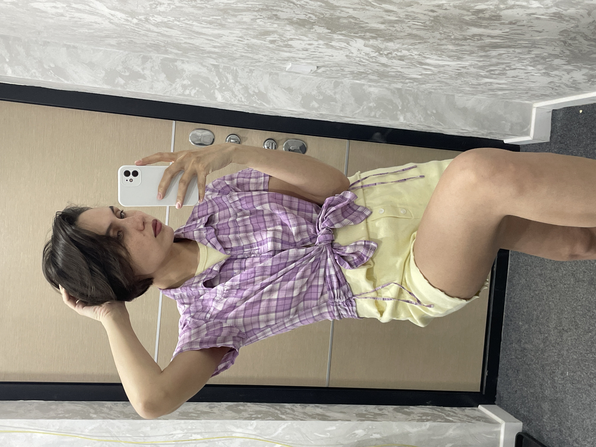 An indoor mirror selfie captures a young individual with short, dark hair and noticeable freckles. The person is wearing a light yellow top underneath a purple and white plaid shirt, which is tied at the waist. They are also wearing light-colored shorts or a similar garment.
The person holds a white smartphone with their right hand, positioned to take a reflection shot in what appears to be a full-length mirror. Their left hand rests gently on the back of their head, amidst their hair. The expression is neutral and focused, seemingly posing for the camera.
The background, visible through the mirror's reflection, consists of a light-colored, possibly wooden or textured, wall or door behind the person. Below this, there is a prominent black frame or molding. Above the frame, a distinct grey and white textured wall with a wavy, marble-like pattern is visible, and below the frame, a grey carpeted floor with a slightly rough texture can be seen. The lighting suggests daytime. The setting is a private indoor space, possibly a home or hotel room in Izhevsk, Russia. No visible text is present in the image.