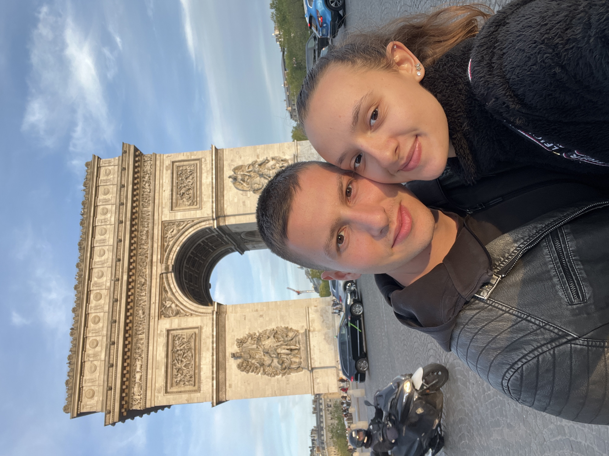 A selfie captures a young couple in front of the iconic Arc de Triomphe in Paris, France. The image is oriented vertically, showing the man and woman in the foreground with the large historical monument directly behind them.
The woman is on the right side of the frame, smiling subtly, with her head gently resting on the man's shoulder. She has long brown hair and is wearing a dark, possibly black, fluffy or fleece-like jacket. Her ear is visible, adorned with several small, intricate piercings, including a flower-shaped one and a small stud. The man, on the left, also smiles faintly and is wearing a black leather jacket over a dark hoodie. Both appear to be enjoying their time together.
Behind the couple, the grand Arc de Triomphe rises prominently against a clear, light blue sky with sparse, wispy clouds. The monument's intricate stone carvings and sculptural details are clearly visible, including relief panels and decorative elements. Through its main archway, a glimpse of the sky and distant elements can be seen. On either side of the arch, parts of the monument's façade show detailed sculptural groups.
The setting indicates an urban environment, likely a tourist location, with a street visible in the mid-ground. Several vehicles, including cars (one blue, one black) and a black motorbike, are parked or moving on the road. Other distant figures can be seen, suggesting a bustling area. The lighting suggests it is daytime, possibly late afternoon, with a soft, warm glow. There is no visible text in the image.