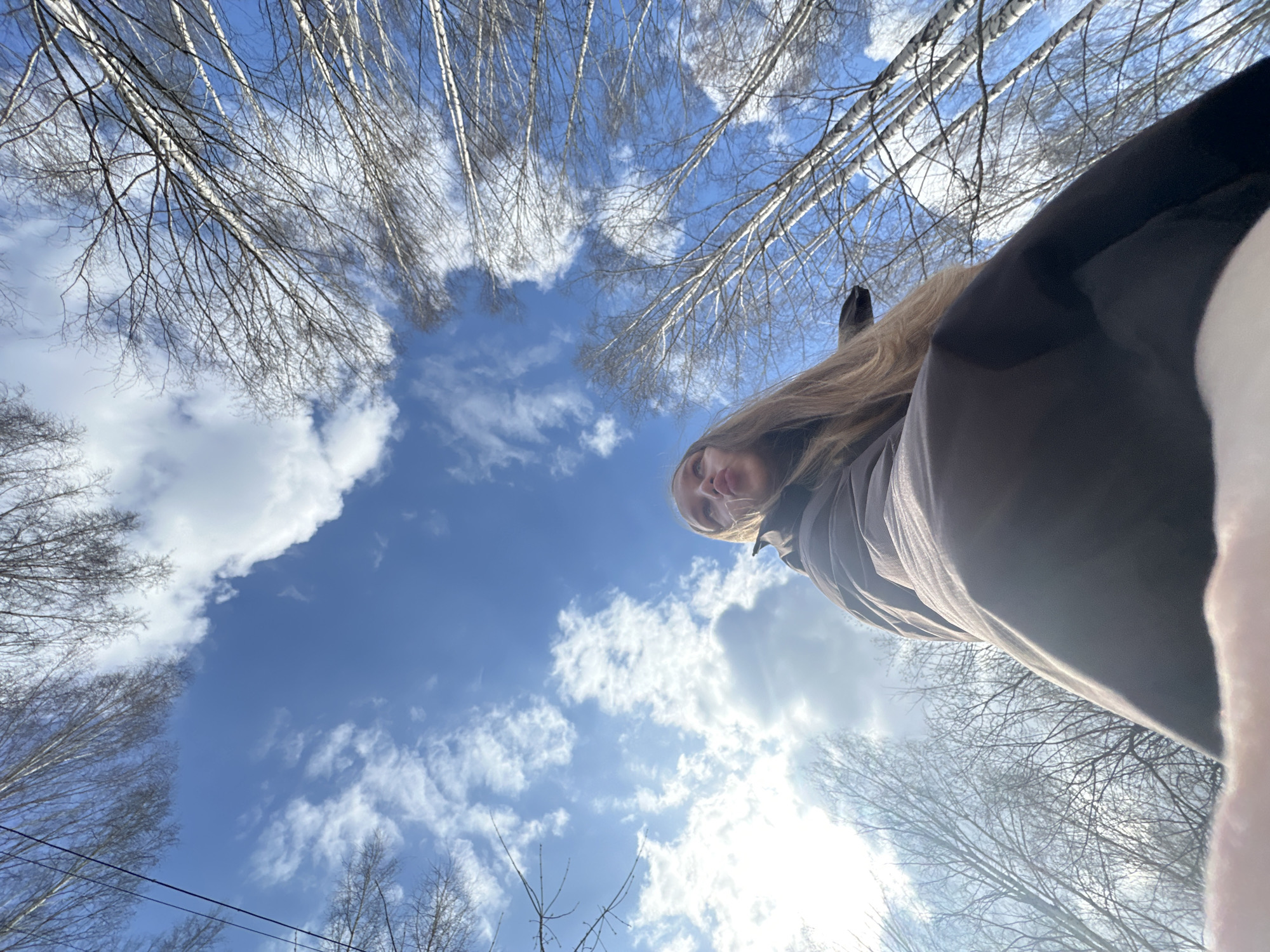A low-angle outdoor shot captures a person, likely female, with long blonde hair, looking down towards the camera. Her face is partially visible in the upper right quadrant of the frame, illuminated by bright sunlight from the lower right. She is wearing a dark-colored jacket. The perspective is looking up from the ground towards the sky and the bare branches of numerous trees, consistent with birch trees, suggesting late winter or early spring.
The background features a clear blue sky dotted with scattered white, fluffy clouds. The sun is prominent, creating a strong glare in the lower right of the image. The trees, devoid of leaves, create intricate patterns against the sky. A thin, dark line, possibly a power line or wire, is faintly visible in the lower left section of the sky. The scene is set outdoors in a forested or park-like area in Chaikovsky, Russia, on a sunny day. No text is visible in the image.