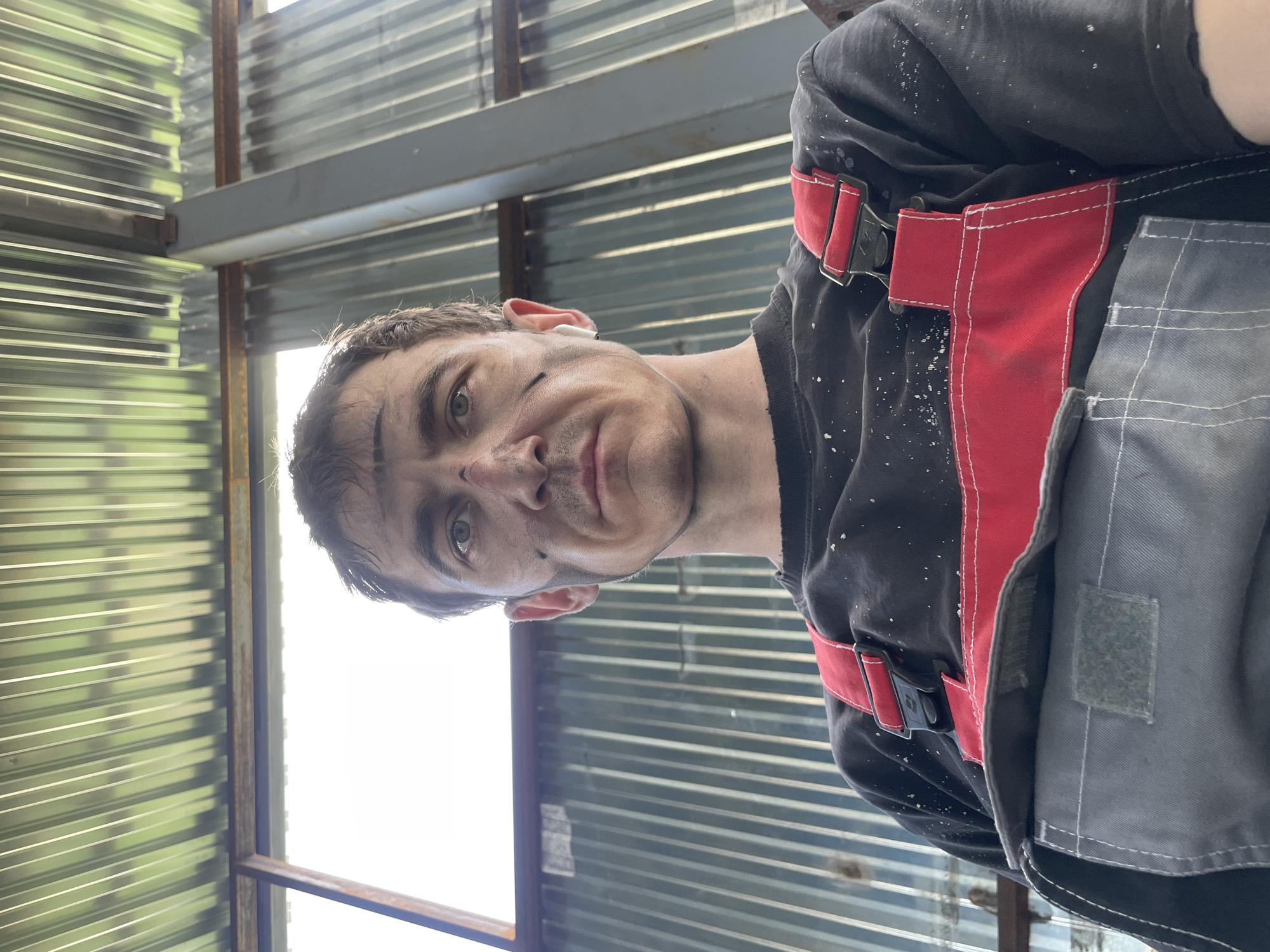 A low-angle, waist-up selfie shows a man with dirt smudges on his face and chest. He is wearing a black t-shirt with white speckles and red and gray overalls. The man has short, brown hair and is looking directly at the camera with a neutral expression. He has a white earbud in his right ear. Behind him, a corrugated metal wall and ceiling are visible, with a large window on the left side of the frame. The overall setting appears to be an industrial or workshop environment. The lighting suggests it is daytime, possibly outdoors or in a well-lit indoor space. The image was taken in Sokury, Russia.