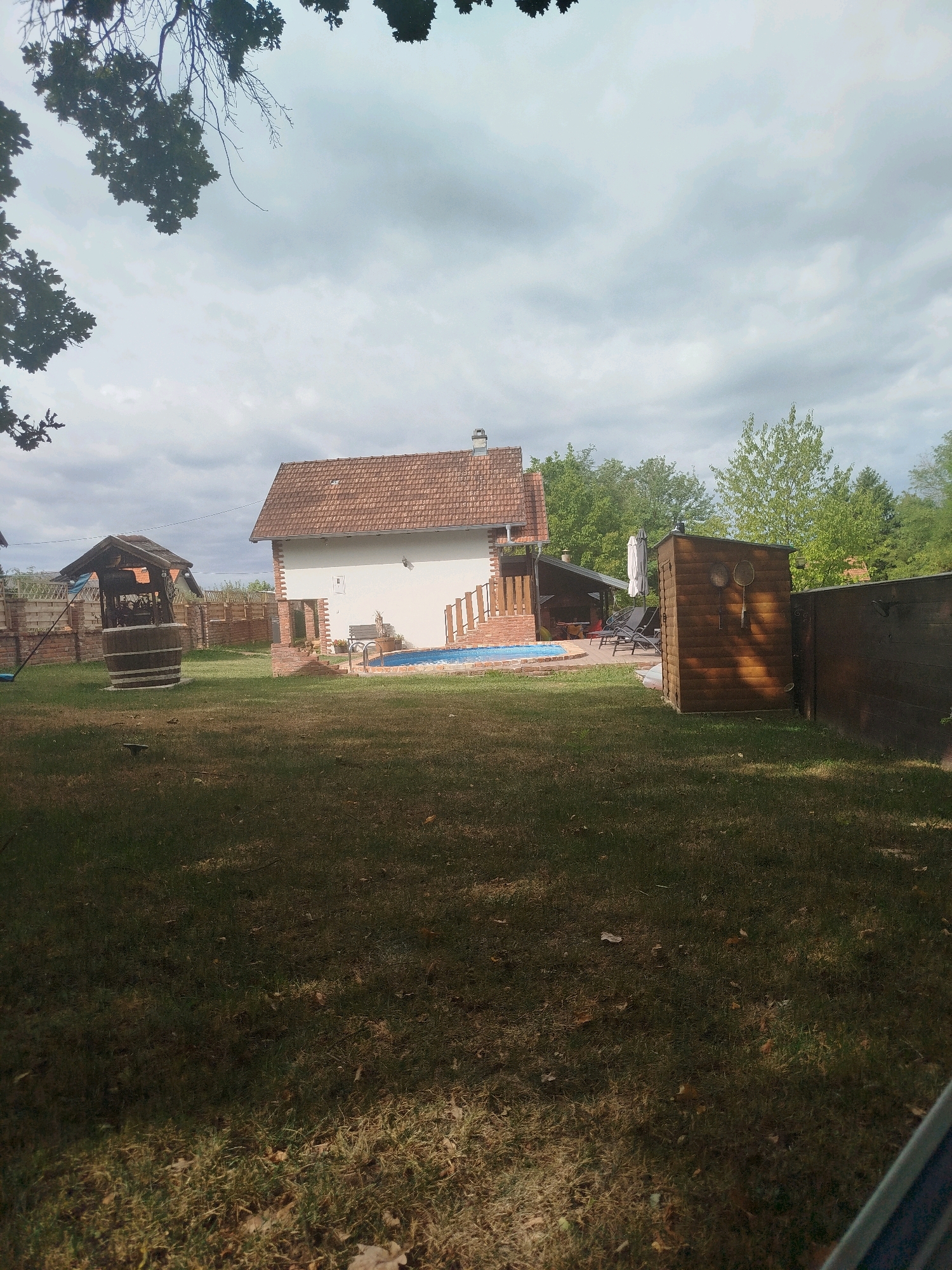A backyard scene in Brckovljani, Croatia, features a white house with a terracotta roof, a blue swimming pool, a wooden shed with badminton rackets hanging on it, and a well with a wooden barrel. The yard is covered in green grass with some fallen leaves. The sky is overcast with dramatic clouds. Trees surround the property, and a wooden fence encloses part of the yard.