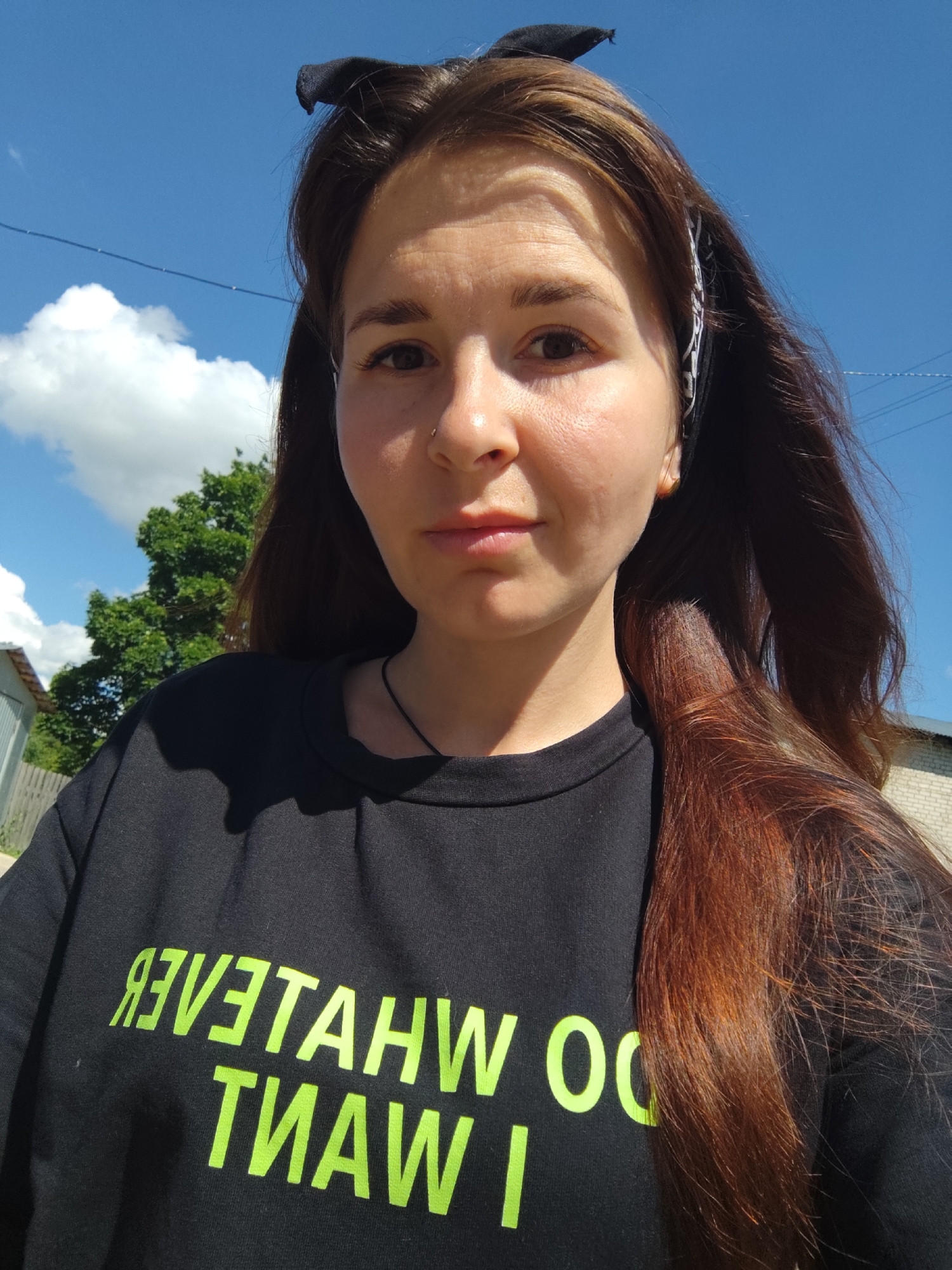 A young woman with long, reddish-brown hair and a black t-shirt with neon green lettering is pictured outdoors. The t-shirt has a reflective inscription that reads "I WANT WHATEVER" in reverse. She is wearing a thin black choker necklace. A black bandana is tied in her hair, protruding upwards like small ears. The background shows a bright blue sky with scattered white clouds, a green tree, and a building that appears to be made of light-colored brick or concrete. The scene appears to be taken on a sunny day. The location is identified as Foky, Russia.
