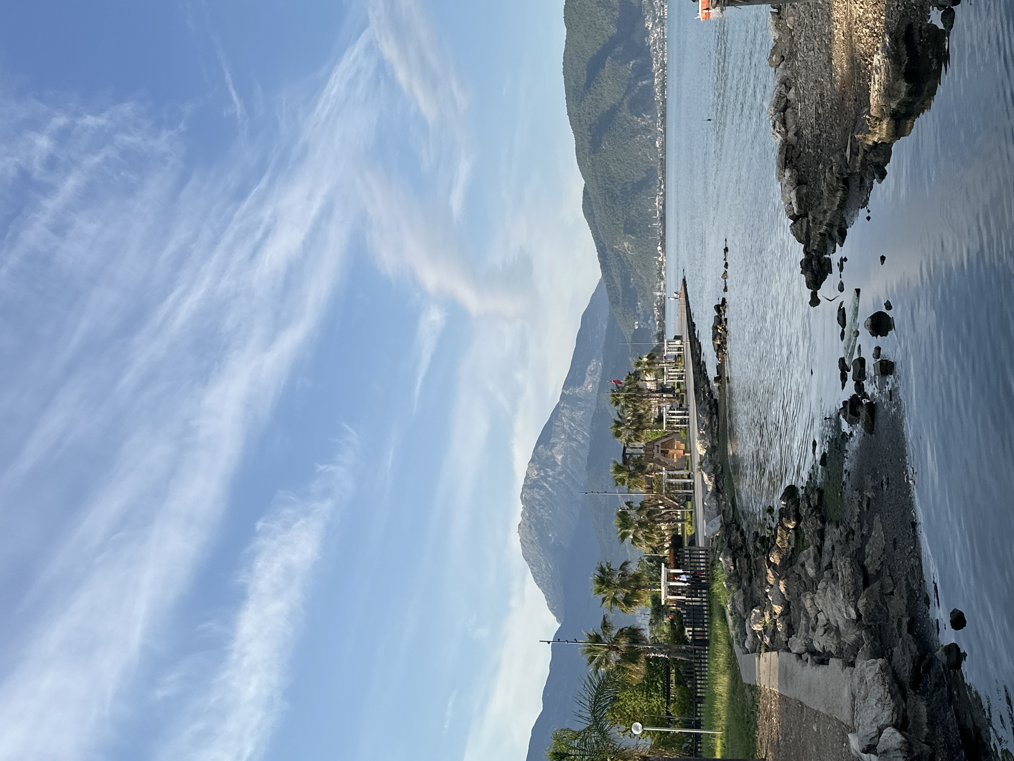 This image captures a serene coastal landscape in Fethiye, Turkey, under a clear blue sky. The sky is adorned with delicate, wispy white cirrus clouds, indicative of fine weather.
Dominating the background are large, verdant mountains, their slopes covered in lush greenery, descending towards the calm, expansive body of water, likely a bay. Along the distant base of these mountains, the shoreline features scattered buildings, suggesting a town or settlement.
In the mid-ground, the tranquil sea reflects the light, showing gentle ripples. On the right side of the foreground, a rugged, rocky breakwater or natural rock formation extends into the water, separating it from a darker, possibly shallower, inlet. To the left of this rocky edge, a concrete path runs parallel to the shore.
The immediate foreground on the left side is lined with a variety of palm trees and other green foliage, indicating a developed promenade or park area. Among the trees and along the path, several establishments with outdoor seating, possibly cafes or restaurants, are visible, including a rustic wooden hut-like structure. Streetlights are sparsely placed along the path. A small red flag can be seen near one of the distant buildings on the left.
A few indistinct figures of people are scattered along the distant shoreline and possibly seated at the foreground establishments, enjoying the pleasant day. The scene conveys a peaceful and relaxing atmosphere, likely captured in the late morning or early afternoon given the bright, even lighting. There is no visible text.