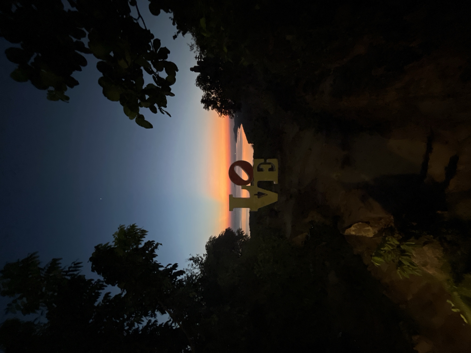 The image depicts a sunset scene in Karimunjawa, Indonesia, featuring large yellow letters spelling out "LOVE" in the foreground, positioned on a rocky path. The letters are illuminated, with the "O" appearing red. Behind the letters, a body of water stretches to the horizon, where the sky transitions from a deep blue at the top to vibrant orange and pink hues near the water's surface, indicative of a sunset. Silhouettes of trees frame the scene on both the left and right sides, with some leaves visible in the upper right portion. In the distance, the faint outlines of islands or landmasses can be seen. The overall lighting suggests it is either dusk or dawn.
