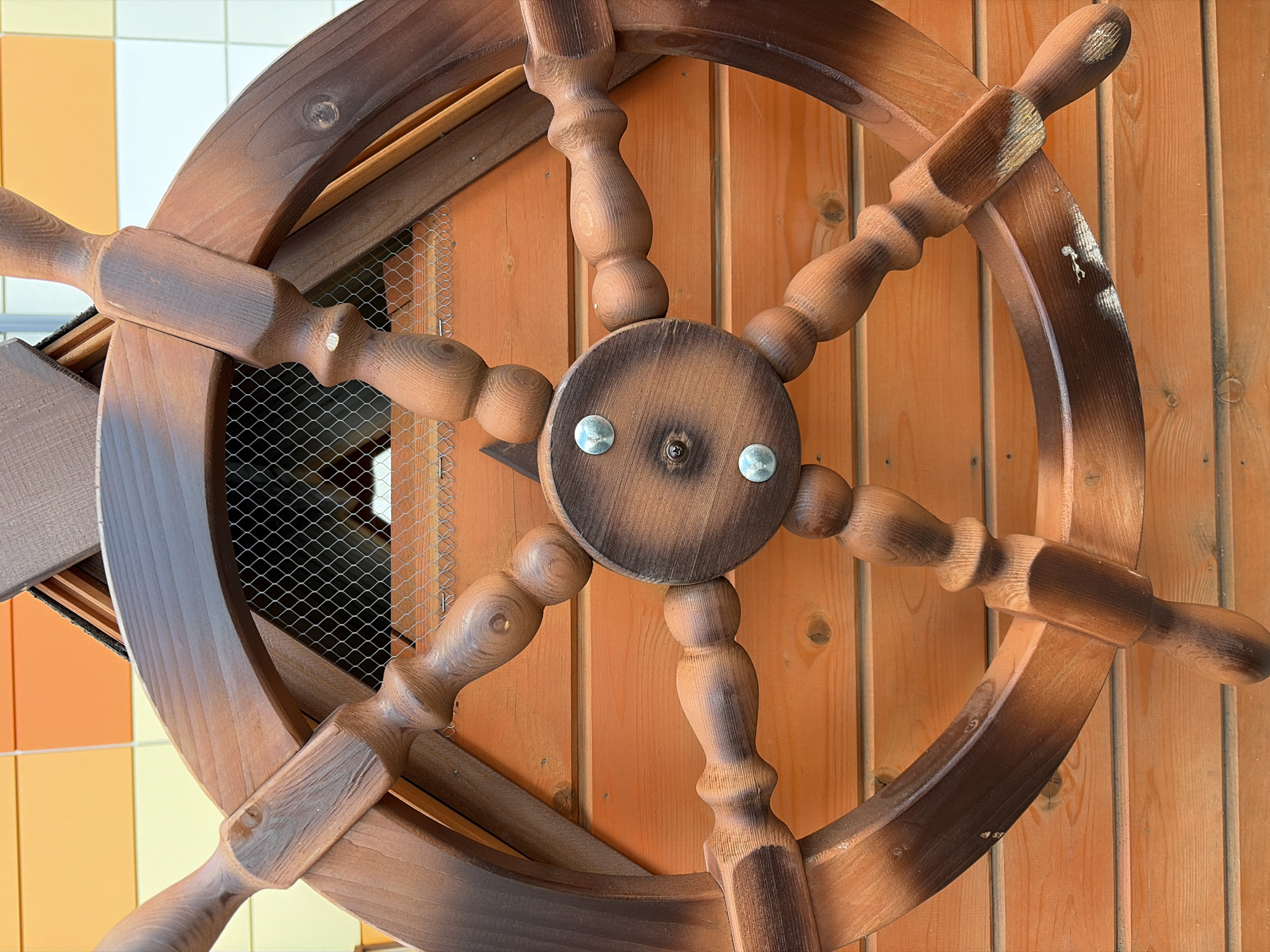 A close-up, eye-level shot shows a large, wooden ship's steering wheel. The wheel is dark brown and has six spokes, each connected to a circular central hub. The spokes are rounded and have decorative bumps and curves. The wheel is mounted on a wooden wall made of horizontal planks, which are a lighter, warmer brown. Behind the steering wheel, a section of wire mesh is visible. In the upper portion of the image, a wall covered with orange and cream-colored tiles can be seen. The scene appears to be an outdoor setting, possibly in a park or playground in the city of Belaya, Russia. There are no people or animals in the image. The lighting suggests it is daytime, with some shadows indicating the direction of the light source. The overall impression is of a decorative or play structure.