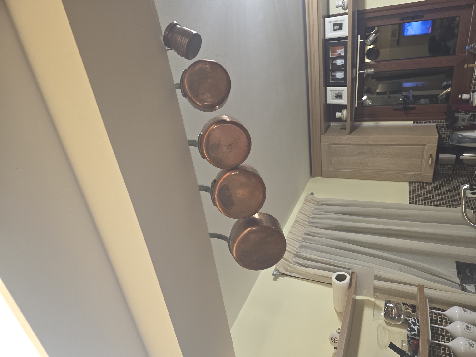 The image shows a kitchen interior. In the foreground, a shelf is visible with several items including a roll of paper towels, a wine glass, and a small decorative bowl. Behind the shelf, a sheer curtain hangs. Above the shelf, a track light with multiple hanging copper pots is suspended from a beam. In the background, wooden cabinets are present, and a window with a wooden frame reflects the interior of the room. On top of the cabinets, framed photographs and decorative vases are displayed. The overall color palette is warm, with dominant tones of cream, beige, and copper. The scene appears to be indoors, likely a residential kitchen, in the Municipal Unit of Asini, Greece.