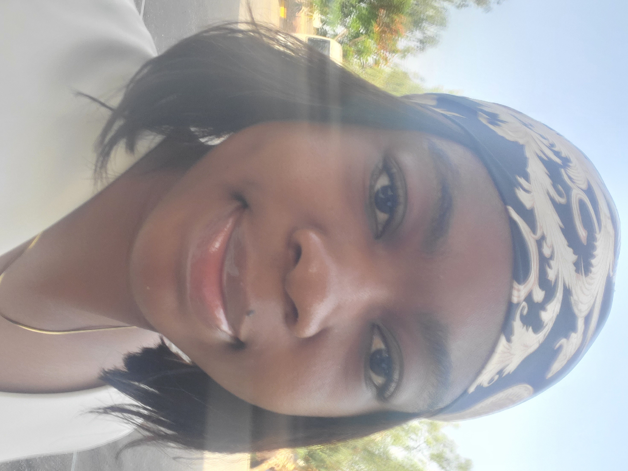 A close-up, selfie-style shot presents a young Black woman with smooth, dark skin. She has a headscarf patterned with white and blue. Her hair is dark, glossy, and short, falling below her ear. A thin, gold necklace is visible at the base of her neck. She is smiling slightly. The background shows green foliage and a hazy light. The location is likely Gwagwada, Nigeria.