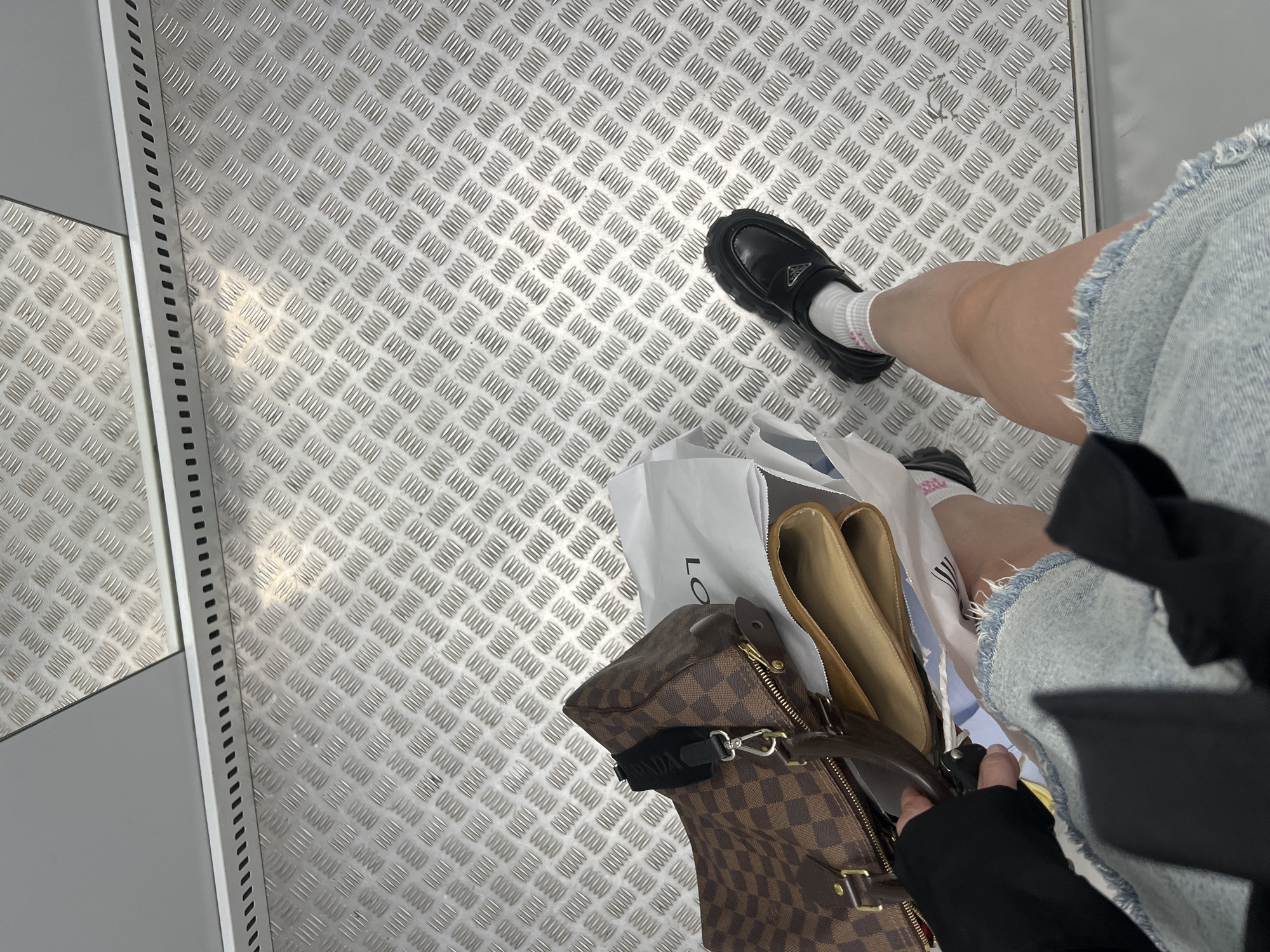 The media file is a top-down, point-of-view shot of a person's legs and their belongings on a textured metal floor. The subjects are a person wearing denim shorts with frayed edges, white socks with pink stripes, and black loafers. They are holding several shopping bags, including a large brown and beige checkered pattern bag with a dark brown strap and metal hardware, and a white paper bag with black text that appears to read "LO". A glimpse of another light brown bag is visible. The scene appears to be indoors, with the metallic floor suggesting a public transport setting, possibly a train or metro station, or a utilitarian industrial space. The lighting is bright, indicating daytime. No specific weather conditions or emotions are discernible from the image. The visible text "LO" on the white bag is the only decipherable writing.