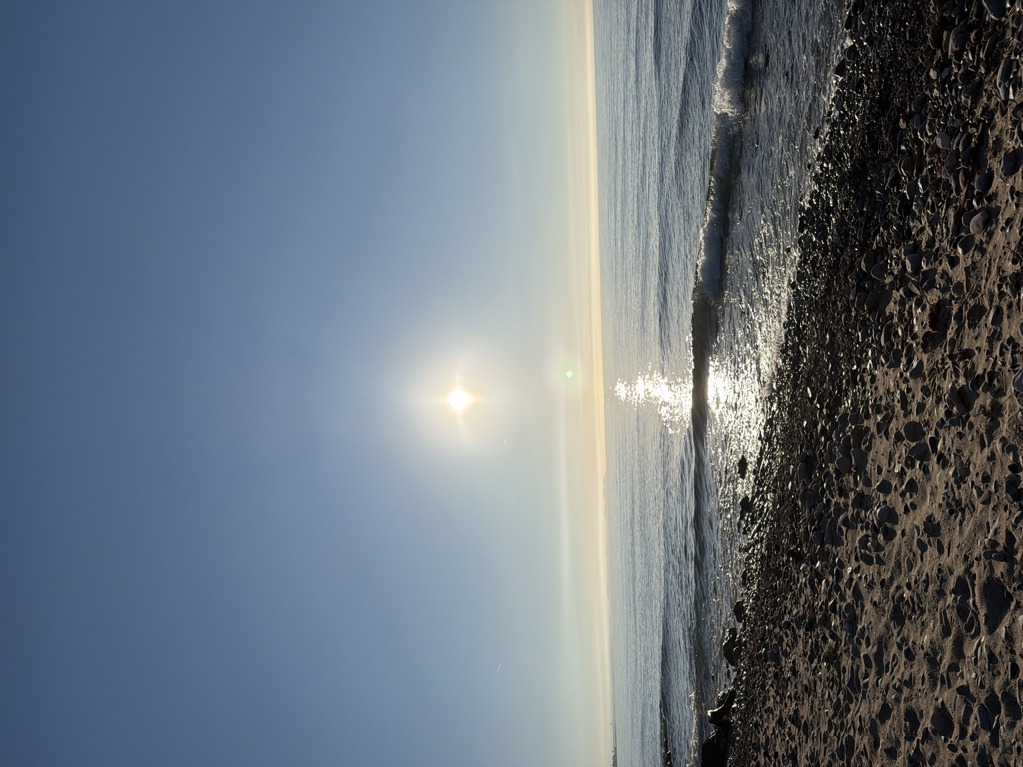 The image depicts a sunny beach scene at Mont-roig del Camp, Spain. The sky is clear blue with the sun prominently positioned. The sea extends to the horizon with some visible waves and sun glare reflecting off the water's surface. The shoreline is pebbly and rocky. No people are visible.
