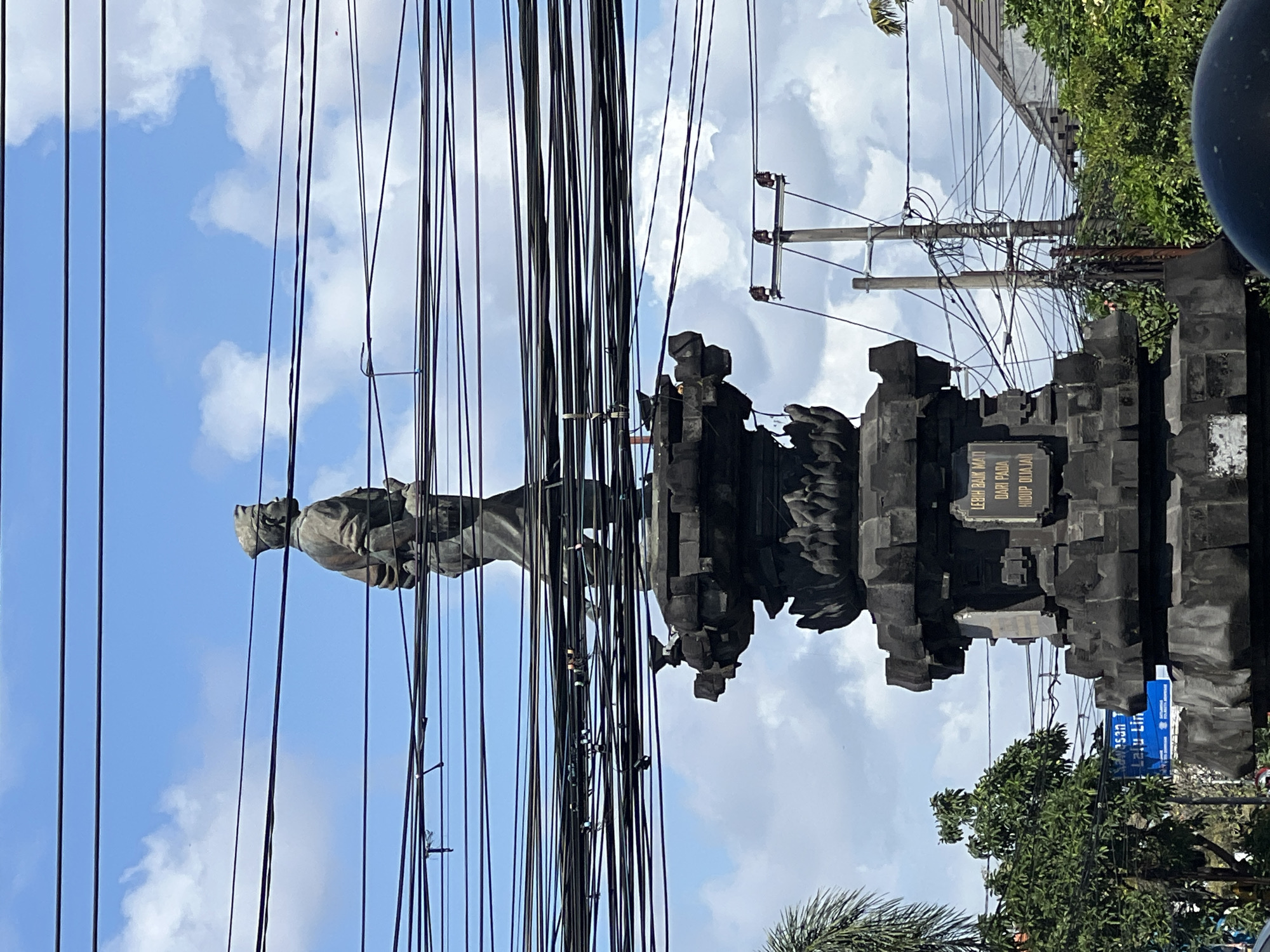 A statue of a man in a hat and uniform stands on a multi-tiered stone pedestal against a bright blue sky with scattered white clouds. Numerous black electrical wires are strung across the frame, creating a dense network in front of and around the statue. The statue is made of dark material, possibly bronze or stone, and depicts the man with his head turned to his right and his arms clasped in front of him. The pedestal is intricately carved with decorative elements. Below the statue, a golden plaque is affixed to the stone, bearing text in Indonesian. In the background, trees and buildings are visible, along with more utility poles and wires. The scene appears to be in an urban or semi-urban environment. The statue is likely a monument, and based on its style and the inscription, it could be related to Indonesian history or local figures. The presence of extensive electrical wiring suggests it is located in an area with significant infrastructure.