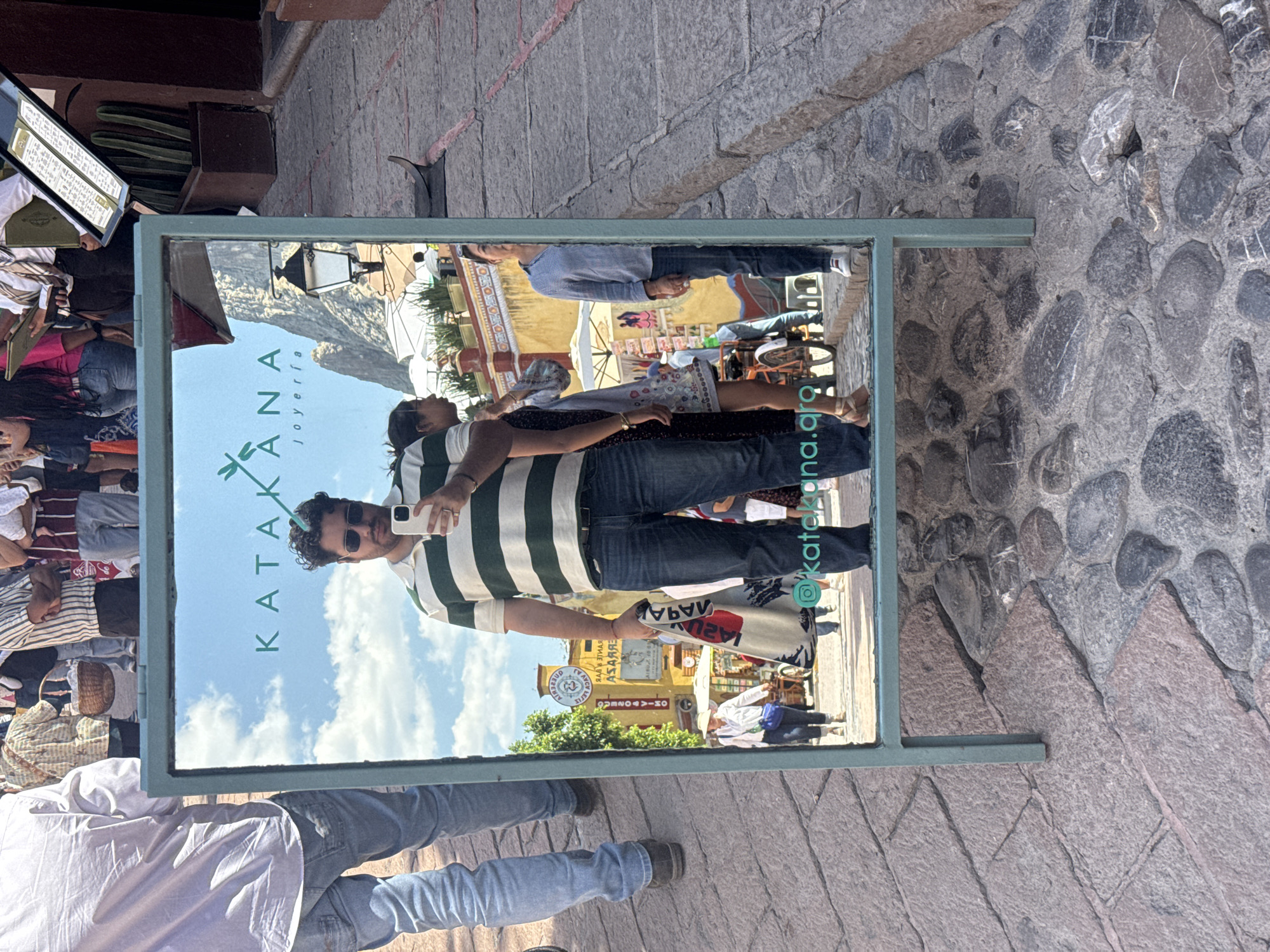 The image shows a mirror positioned on a cobblestone street in Bernal, Mexico, reflecting the surroundings and people. The reflection includes the storefront of "KATAKANA joyería" and other nearby shops. People are visible both in the reflection and directly in front of the mirror, including a man taking a photo of himself in the mirror, wearing a striped shirt and sunglasses. The environment outside the reflection consists of the cobblestone street, stone walls, and other pedestrians.