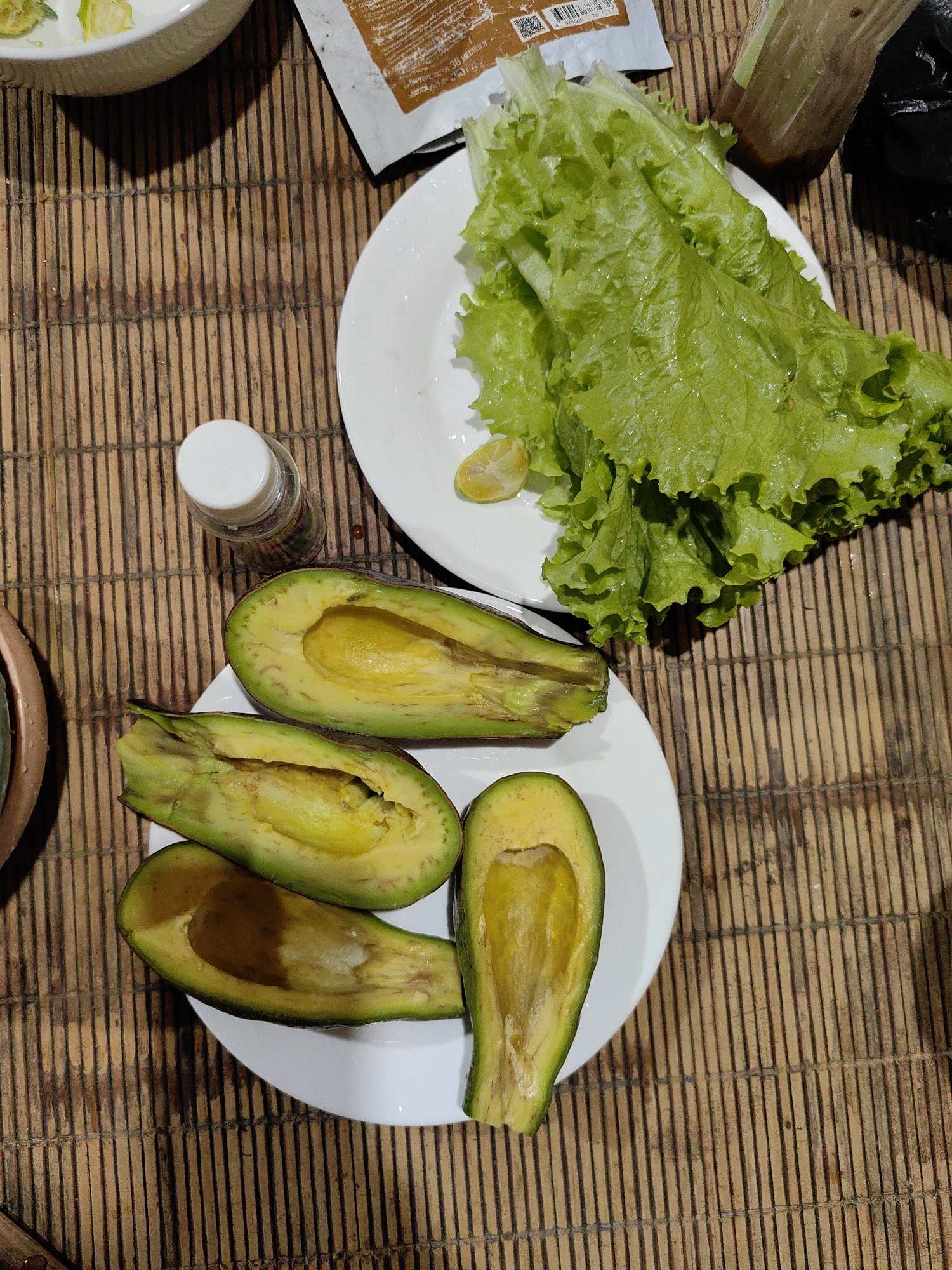 The image depicts a tabletop scene in Xã Đàm Thủy, Vietnam, featuring culinary elements arranged on a bamboo mat. A white plate holds several halved avocados, their green skins slightly darkened in places, revealing the yellow flesh inside. Another white plate is piled with fresh green lettuce leaves and a slice of citrus fruit. A small, transparent bottle with a white lid is positioned near the avocado plate. A white bowl, partially visible at the top left, contains food mixed with what appears to be lime. A package lies near the lettuce plate, and there is a container of sauce and a black object. A portion of a dish is visible at the bottom left.