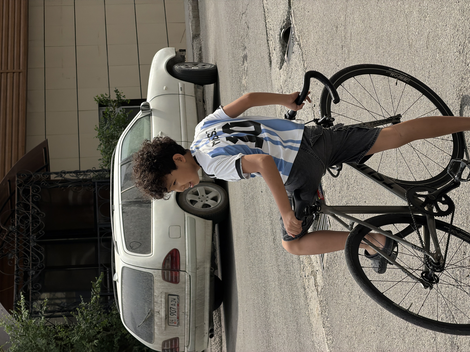 A young boy wearing an Argentine soccer jersey with the number 10 and "MESSI" on the back, stands with his foot on the seat of a bicycle. He is looking to his right, with a slight smile. The boy has curly dark hair and is wearing denim shorts and dark crocs. He is positioned on a paved street in front of a white station wagon. The car, which appears to be a Mercedes-Benz E-Class, is parked on the side of the road. Behind the car, there is a building with a modern facade and an ornate, dark-colored entrance. Green foliage is visible on the left side of the image, near the building. The overall scene suggests a casual, outdoor setting in a residential or urban area.