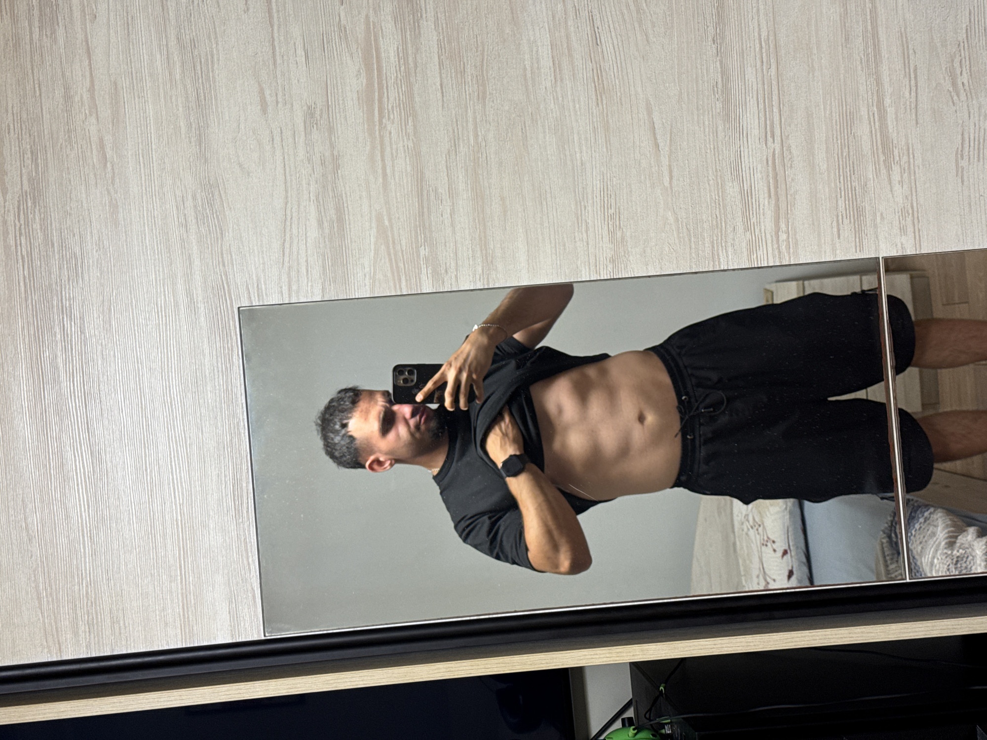 A man is standing in front of a mirror, taking a selfie. He is wearing a black t-shirt and black shorts. His t-shirt is lifted to reveal his abs. He has short, dark hair and a beard. He is wearing a watch on his left wrist. The mirror is reflecting a bed in the background. The man appears to be in a bedroom. The image is taken in Shumen, Bulgaria.