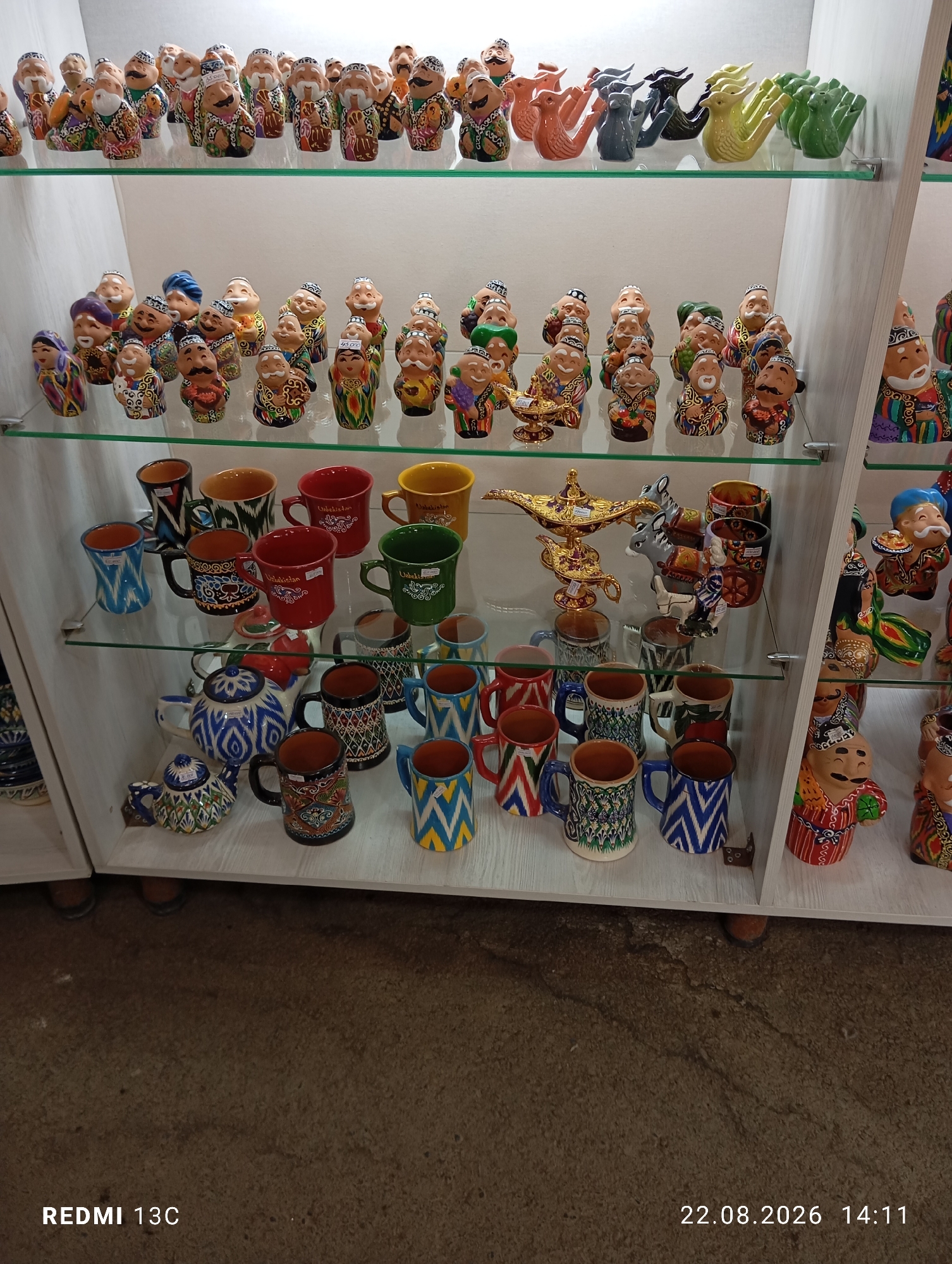 In Amirsay, Uzbekistan, a display shelf showcases a variety of ceramic handicrafts. The top shelf features a collection of small, brightly painted figures resembling people and whimsical creatures like dragons and birds in various colors such as orange, grey, yellow, and green. Below, the second shelf is filled with rows of similar miniature ceramic figures, predominantly men with mustaches and distinct traditional clothing, and a golden lamp. The third shelf displays a selection of ceramic mugs and teapots with intricate patterns. Mugs in red, blue, yellow, green, and patterned designs are visible, along with a blue and white patterned teapot and a smaller decorative teapot. The bottom shelf continues with more patterned mugs in various styles and colors. The overall environment appears to be a shop or market stall, with a concrete floor visible below the shelves.