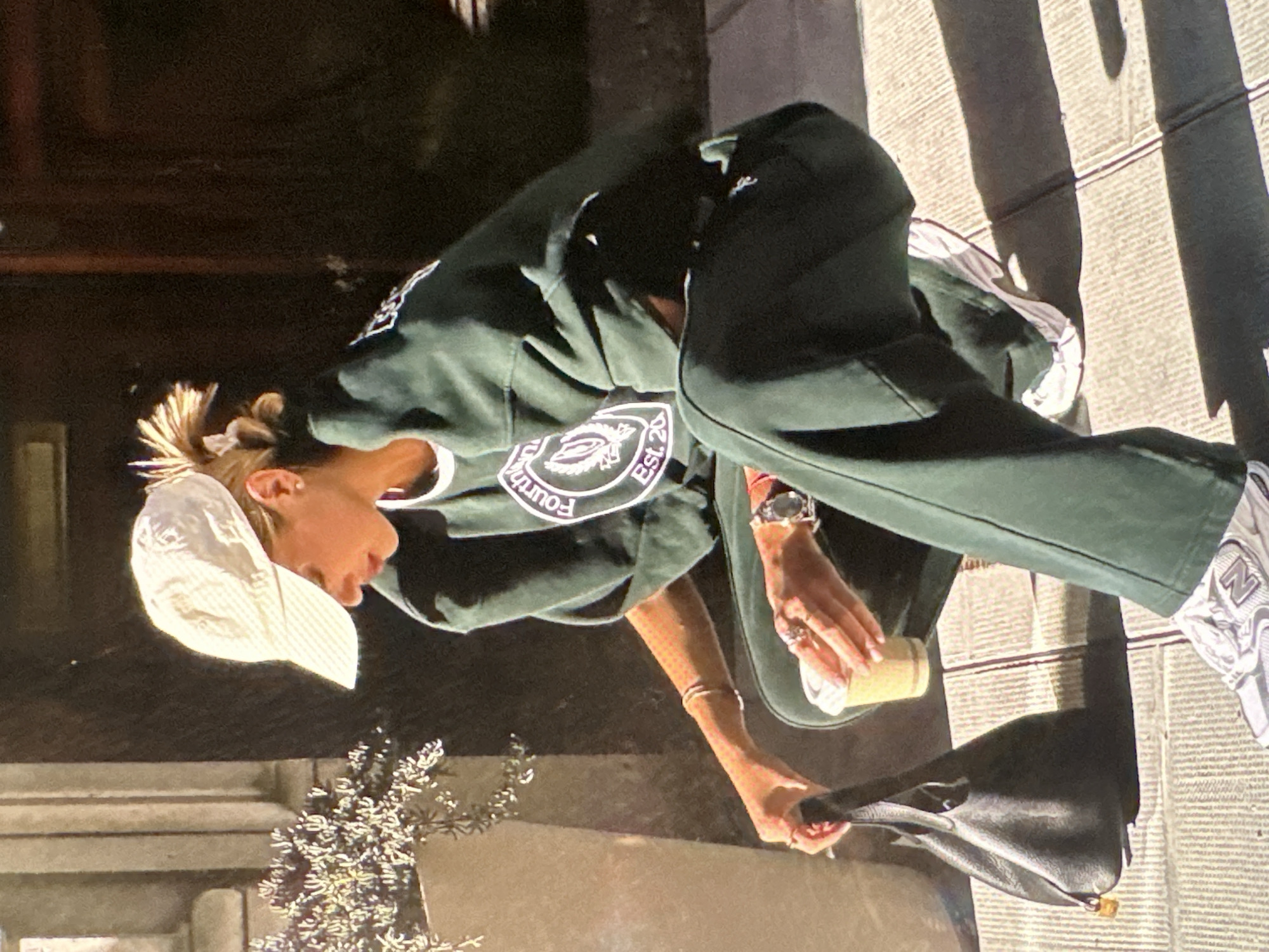 A young woman is standing on a sidewalk next to a building. She is wearing a white baseball cap, a dark green sweatshirt with a circular logo, dark green pants with a white stripe, and white and grey sneakers. She is holding a coffee cup in one hand and a black leather handbag in the other. There is a small bush or plant next to her. The sidewalk is made of concrete slabs with lines creating a grid pattern. The building behind her appears to be made of stone. It is daytime, and the light suggests it may be sunny. The location cannot be determined as Yerevan, Armenia based solely on visual data.