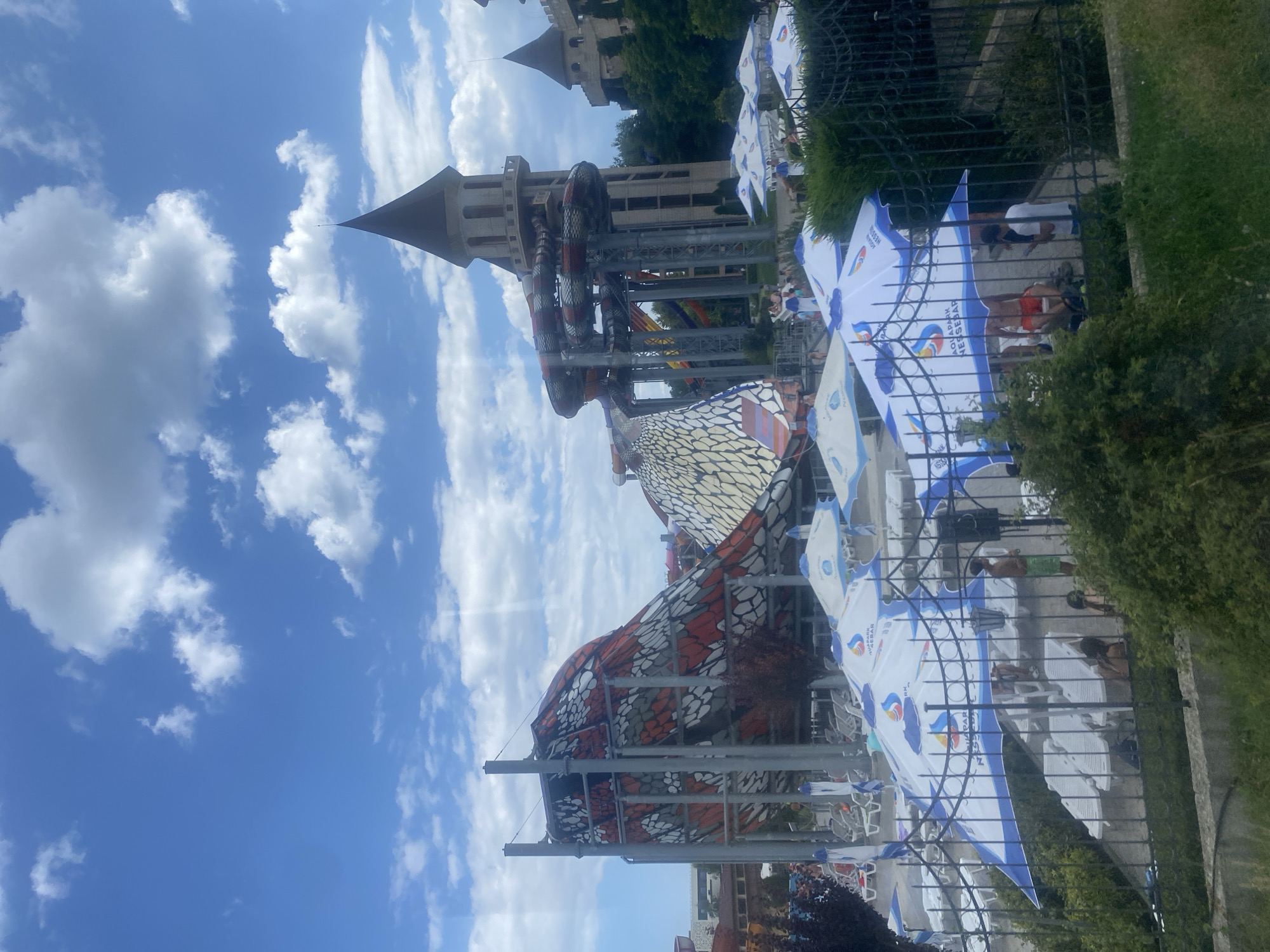 The image shows a view of a water park, likely Aquapark Nessebar, in the vicinity of Ravda, Bulgaria. The water park features prominent attractions, including a large, colorful water slide structure with a patterned design resembling scales, and a towering castle-like building with multiple slides descending from it. In the foreground, white lounge chairs and blue and white umbrellas are set up. Several people are visible, some relaxing on lounge chairs, others walking or standing under the umbrellas. The sky is blue with scattered white clouds. The overall scene depicts a sunny day at a recreational water park.