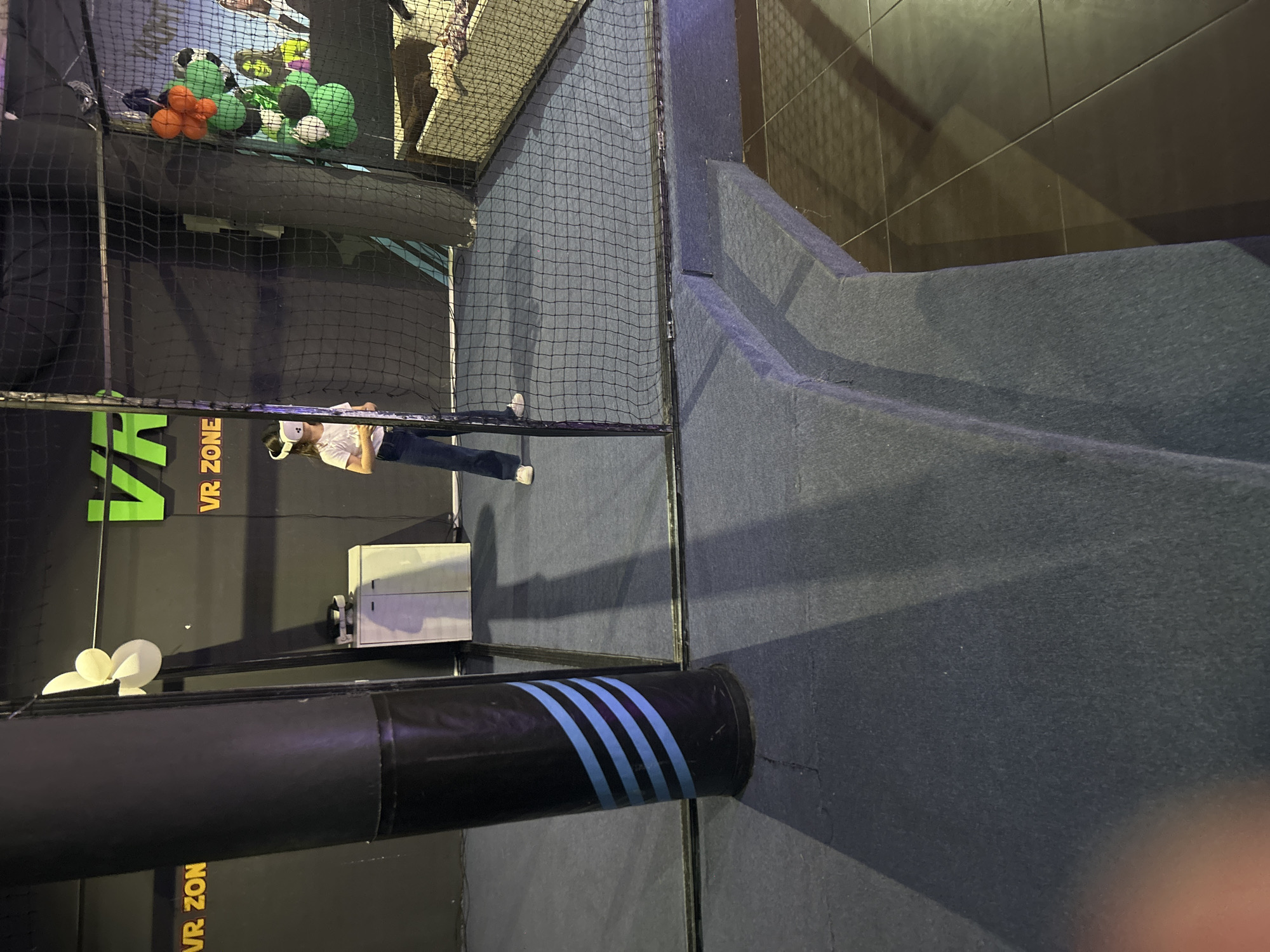 A person wearing a VR headset stands inside a netted enclosure at a VR Zone. The VR Zone is located in an indoor play area with padded flooring and walls. Green and orange ball-shaped objects are above the enclosure. A black cylindrical object with blue stripes is in the foreground. The location is Burgas, Bulgaria.