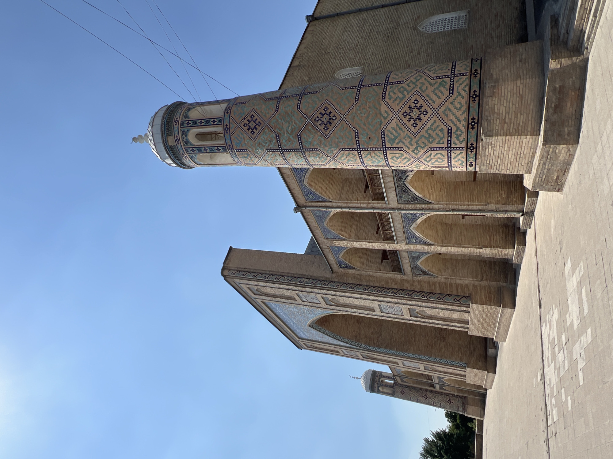 The image depicts a minaret and a section of a building adorned with intricate blue and white geometric tilework. The minaret, a tall cylindrical tower, is decorated with repeating diamond patterns and bands of smaller decorative elements. Its top features a light-colored, ornate finial. Adjacent to the minaret, a building with arched doorways and windows showcases similar tilework along its facade, creating a sense of depth and architectural grandeur. The construction appears to be primarily brick, with light-colored stone accents.
The scene is set outdoors under a clear, bright blue sky, suggesting a sunny day. The sun's angle indicates it is likely daytime, possibly morning or afternoon. The perspective is from a low angle, looking upwards towards the minaret and the building. The ground in the foreground is paved with large, light-colored stones, some of which are stained or discolored, hinting at age or extensive use. The overall impression is one of historical and religious architecture, likely a mosque or a madrasah, given the presence of the minaret. The clean sky and well-preserved tilework suggest the location is a well-maintained historical site in Tashkent, Uzbekistan. No people are visible in the frame, and there is no visible text.