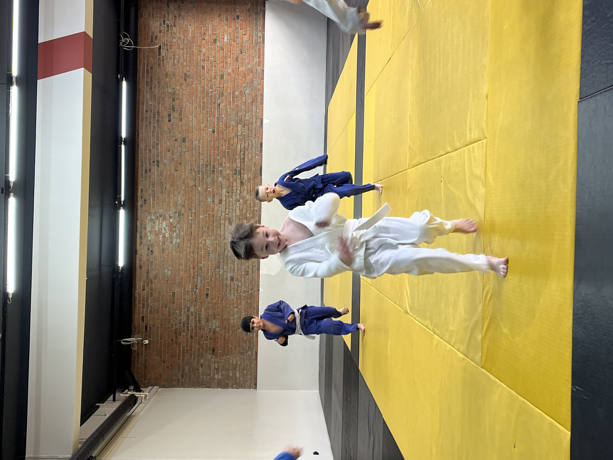 This image captures an indoor scene within a martial arts training facility, likely in Krasnoyarsk, Russia. The perspective is rotated 90 degrees counter-clockwise, but describing it as if correctly oriented: the floor is covered predominantly with bright yellow padded mats, with some black mats visible along one side, characteristic of a judo or combat sports dojo.
The main subject is a young boy in the foreground, wearing a white judogi (judo uniform) with a white belt. He is smiling brightly and looking directly at the camera, suggesting a happy and engaging moment during or after a practice session. His bare feet are visible on the yellow mat.
In the mid-ground, slightly behind and to the left of the central boy, another boy in a blue judogi with a white belt is standing on the yellow mats. His expression is less clear, but he appears focused. Partially visible in the background and foreground are other individuals, also dressed in white or blue judogis, indicating multiple participants in the activity.
The background features a rustic red brick wall on the right, contrasting with a clean white wall or pillar on the left. A distinctive white pillar with a reddish-brown diagonal stripe is also visible on the far left. Several long, vertical fluorescent tube lights are mounted along dark structural elements, providing bright illumination to the training area. Wires and cables are visible near some of the lights and walls.
The scene conveys an atmosphere of active training or a break during a martial arts class, with the central boy's smile highlighting a positive and energetic mood. No specific time of day or weather conditions are discernible due to the indoor setting and artificial lighting. There is no visible text in the image.