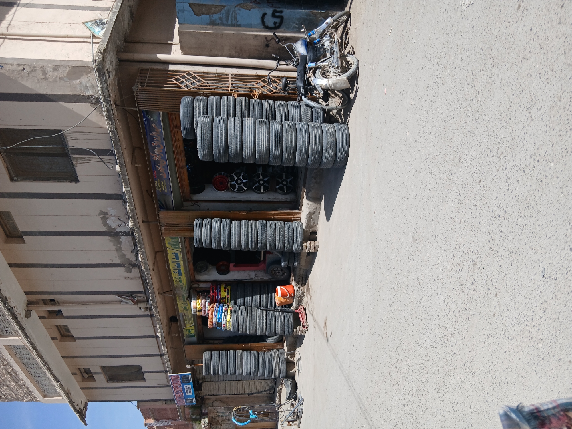 The image shows a tire shop in Quetta City Tehsil, Pakistan. Stacks of tires line the front of the shop. A motorcycle is parked near the tires. The shop is located along a paved road.