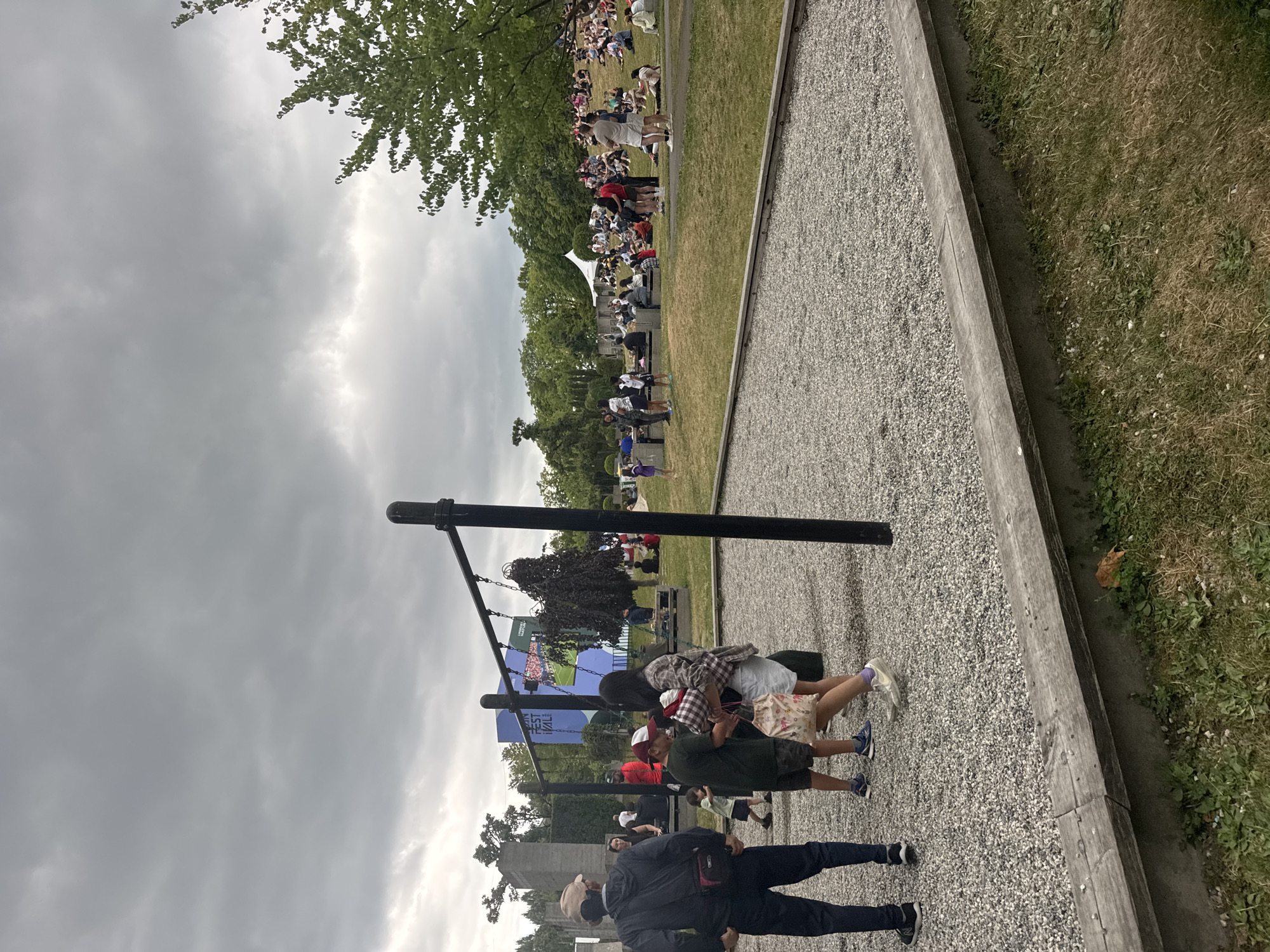 The image depicts a gathering of people outdoors, possibly at an event or festival, under a cloudy sky. In the foreground, a gravel pit with a wooden border suggests a play area or recreational space. A swing set structure stands prominently in the middle ground. Several people, including children and adults, are visible interacting in this area. Further in the background, a large crowd is seated and standing on a grassy slope, facing a stage or screen where a projection or performance is taking place, indicated by a large blue banner with text. Trees and vegetation surround the area, and architectural structures, possibly ruins or art installations, are visible on the left. The presence of the banner "VANCOUVER FESTIVAL" suggests the location is in Vancouver, Canada. The overall scene conveys a lively, community-oriented atmosphere with a focus on outdoor entertainment and public gathering.