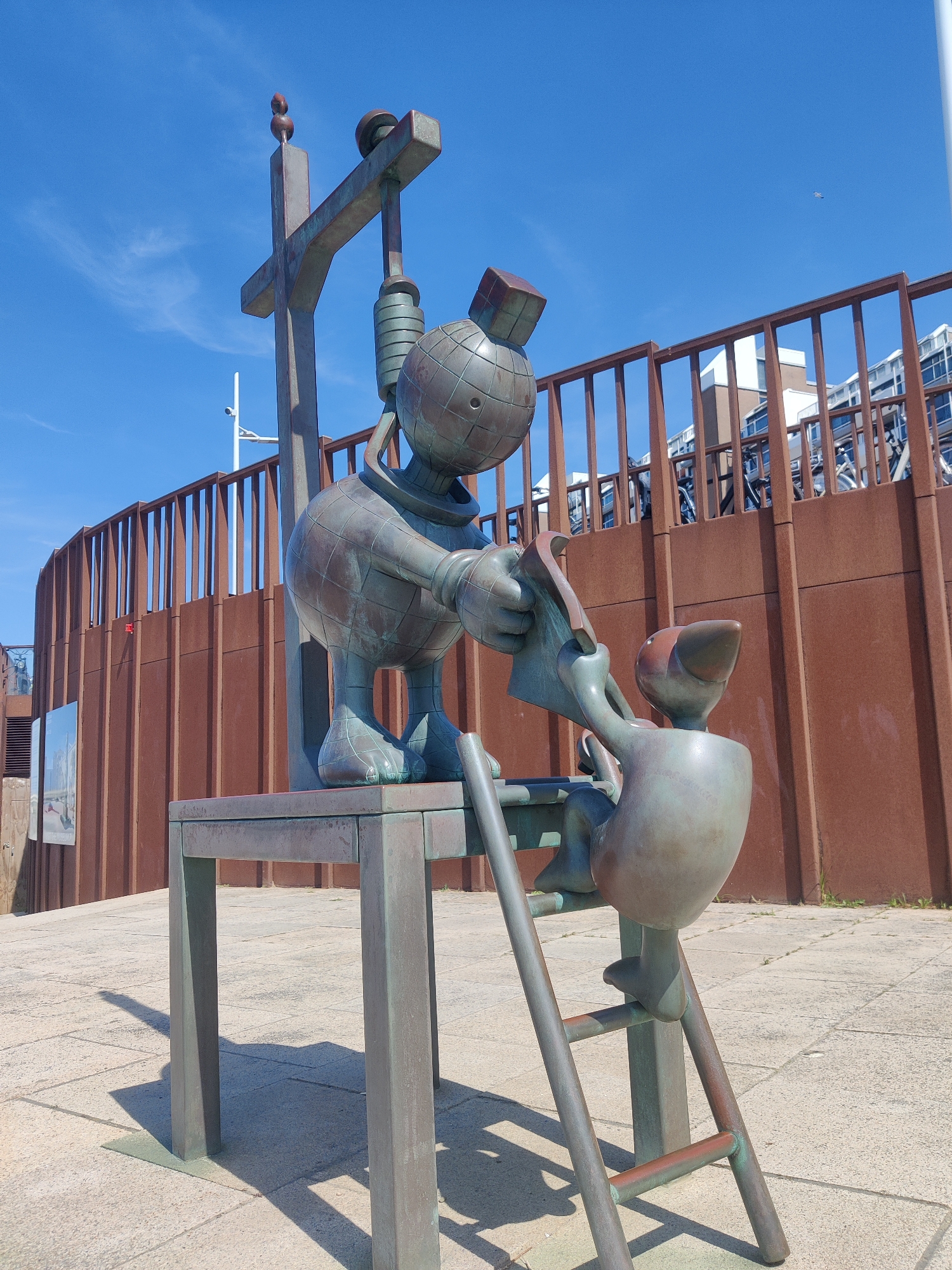This is a low-angle, eye-level, full shot of a bronze sculpture on a sunny day in The Hague, Netherlands. The sculpture depicts two cartoonish figures on a table with a ladder. The larger figure, on the left, is seated on a table, wearing a globe-like body with a cubic head and holding a large piece of paper. The smaller figure, on the right, is climbing a ladder, reaching towards the larger figure. The sculpture is made of weathered bronze, showing signs of green patina. In the background, a tall, rust-colored fence surrounds buildings. The sky is clear blue with a few wispy clouds. The ground is paved with light-colored stone slabs. The sculpture is located at the Hague University of Applied Sciences campus.
